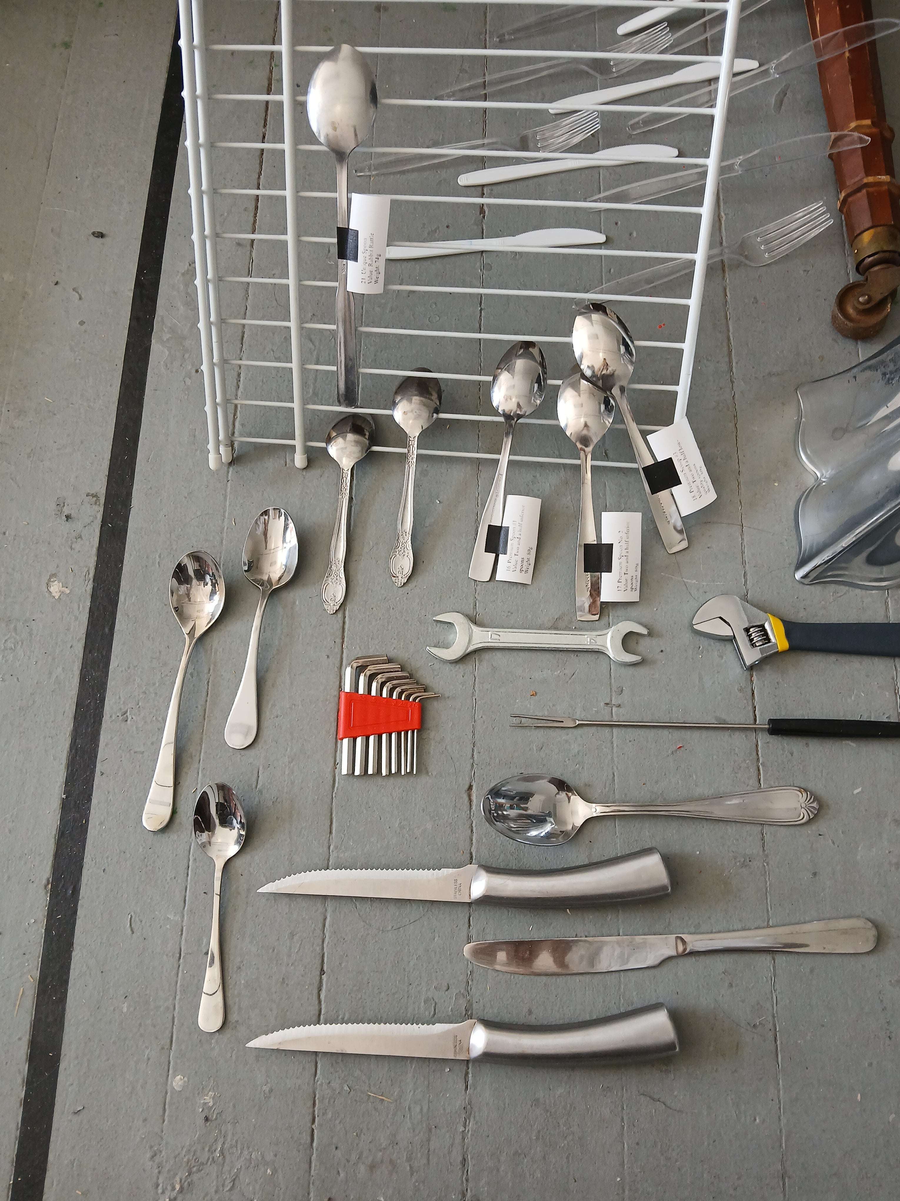 Assorted spoons, knives, and utensils arranged on a gray tiled floor beside a white drying rack, with plastic cutlery, hex keys, a wrench, and an adjustable spanner, some with white labels.