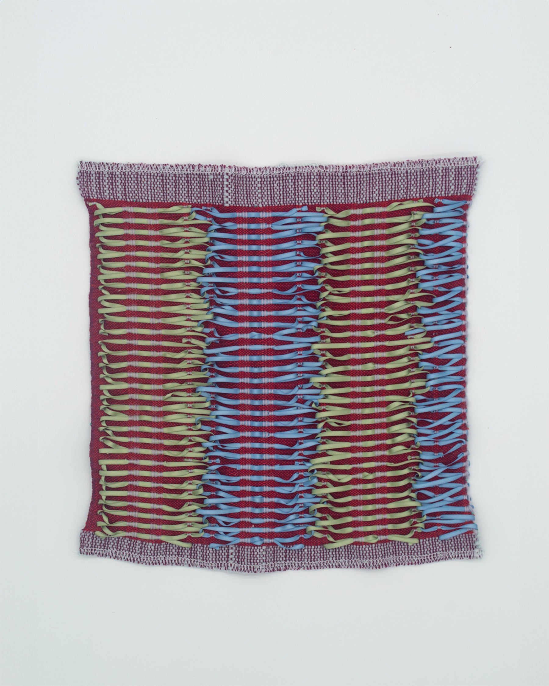 A maroon woven fabric with warm green and baby blue ribbons tied onto the surface. The ribbons run vertically, interlocking in a repeating pattern reminiscent of corset lacing