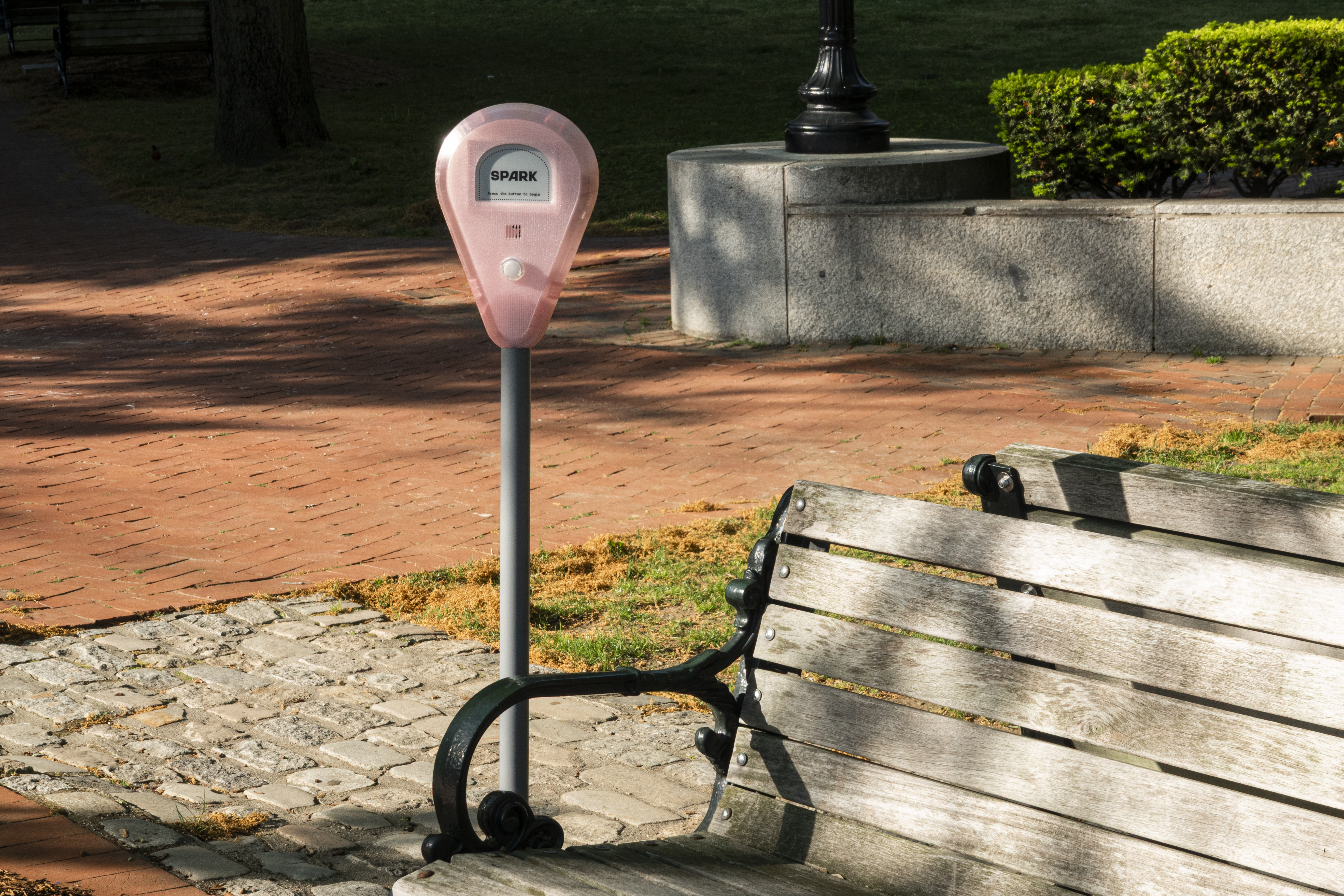 Spark device installed beside a park bench on a brick walkway, with trees, grass, and sunlight in the background.