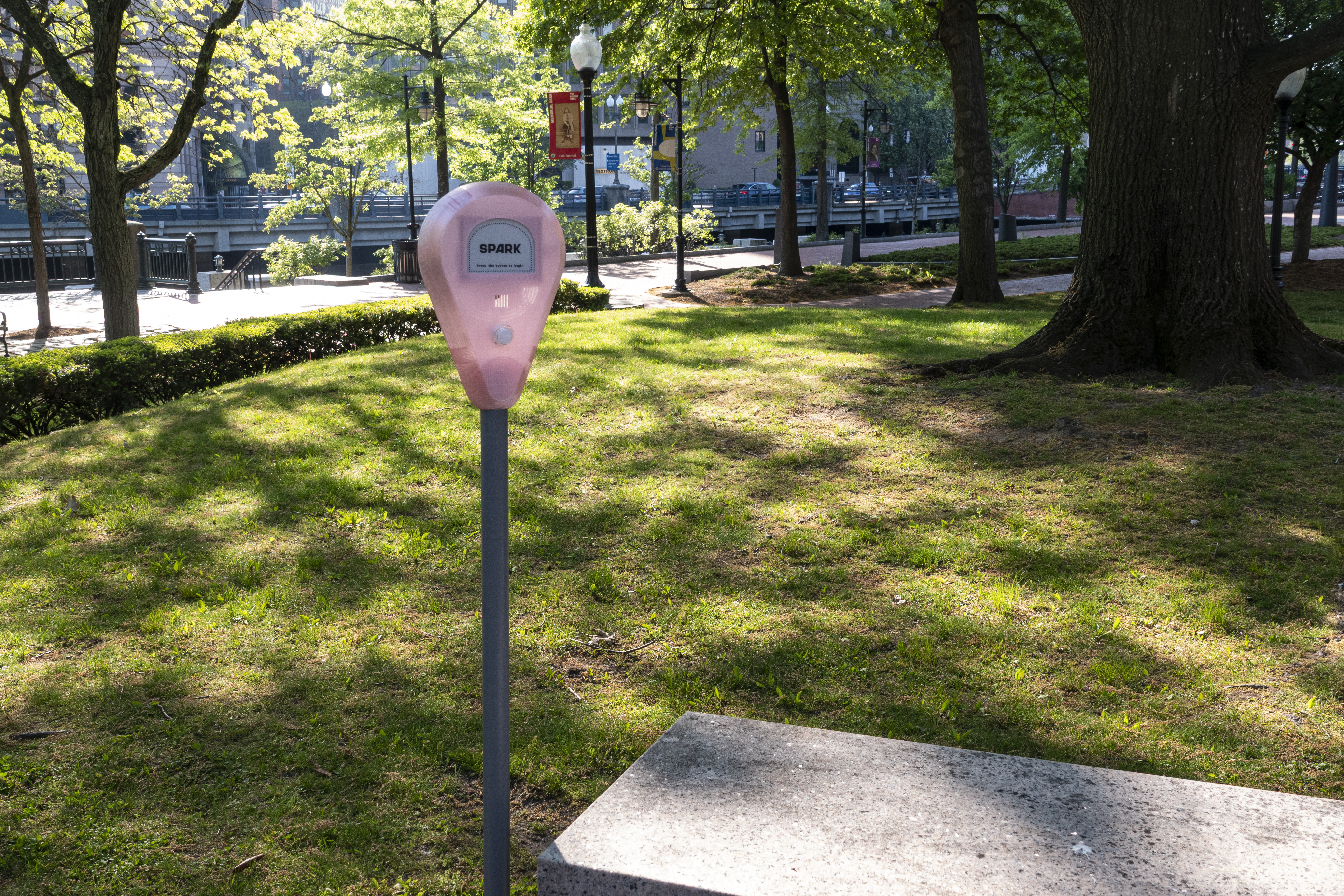 Spark device standing on grass near the river, surrounded by trees, shadows, and city park scenery.