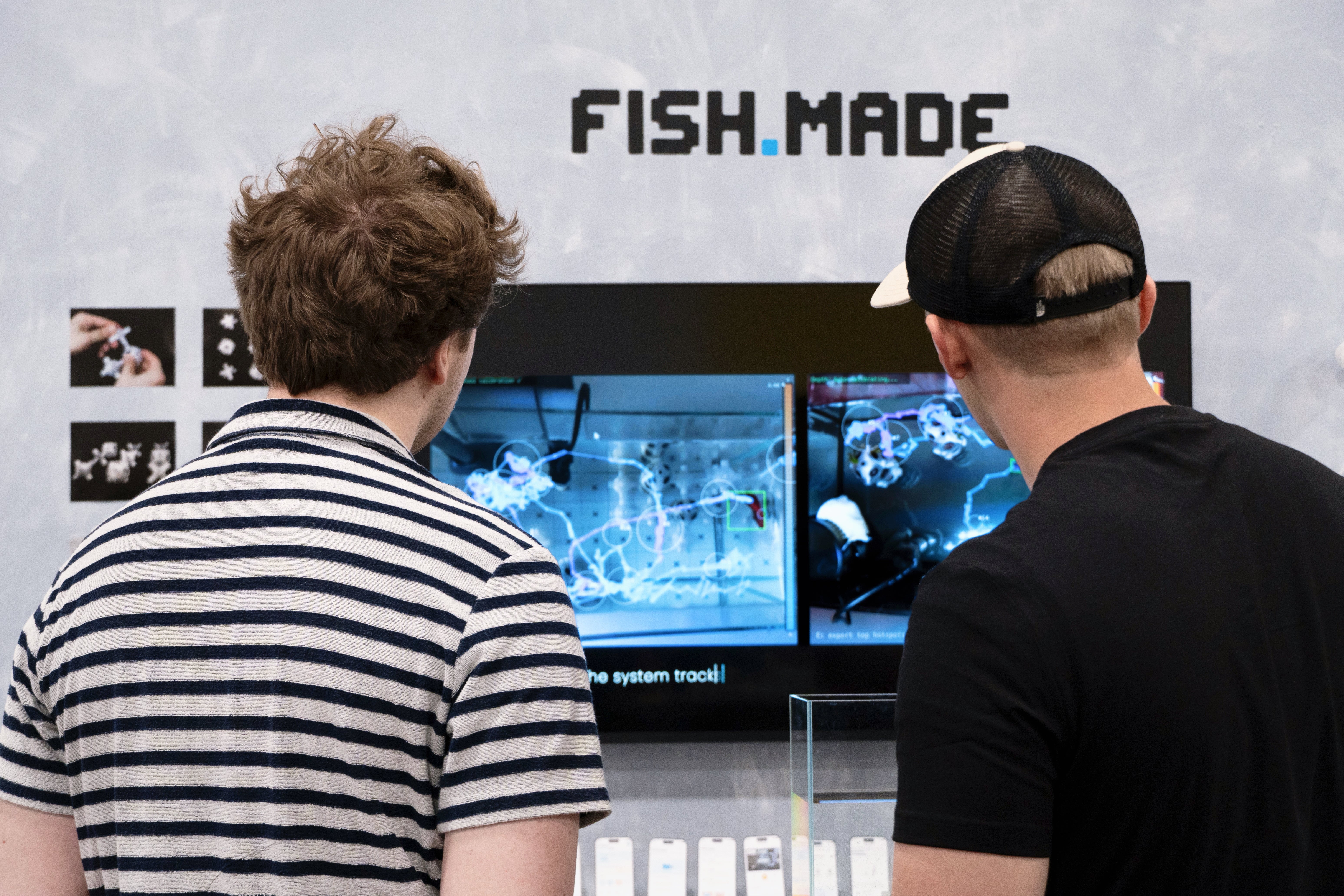 Two visitors view the FISH.MADE installation, which shows fish movement tracking visuals on a screen alongside the project logo and physical prototype display.