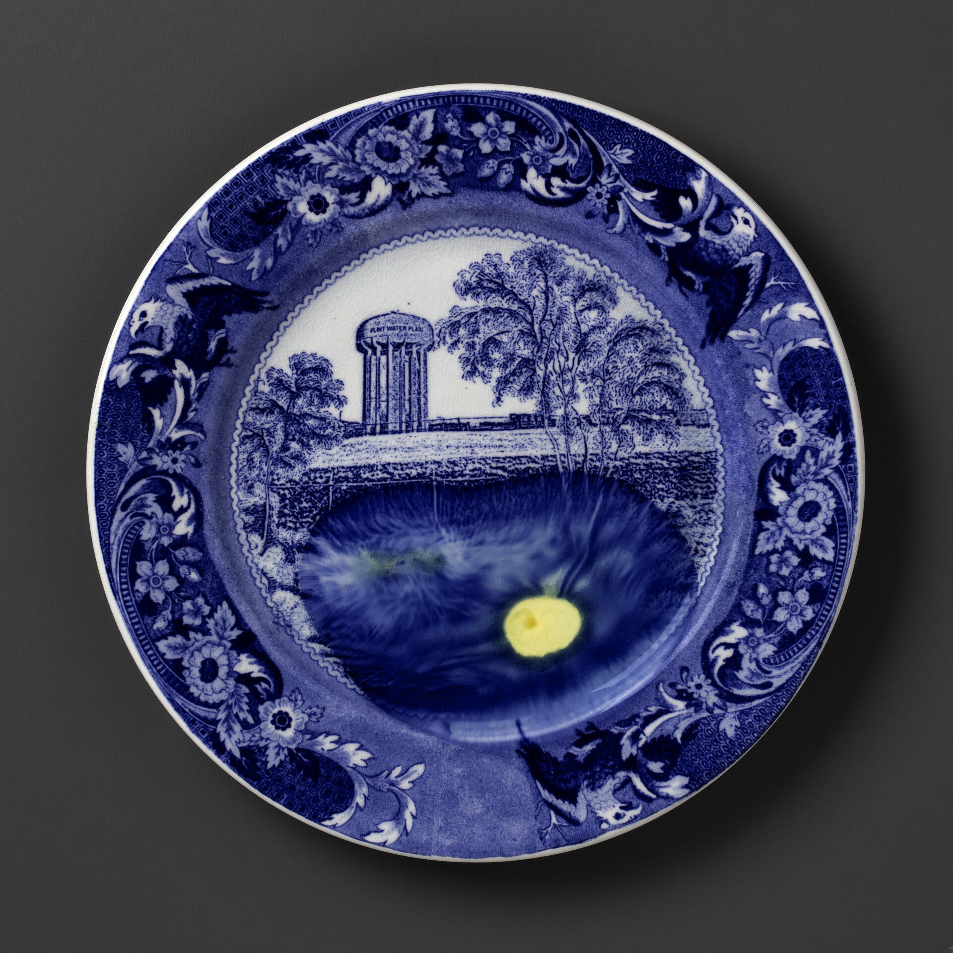 blue and white transferware plate depicting trees and a water tower labeled Flint, with lead melted onto the plate