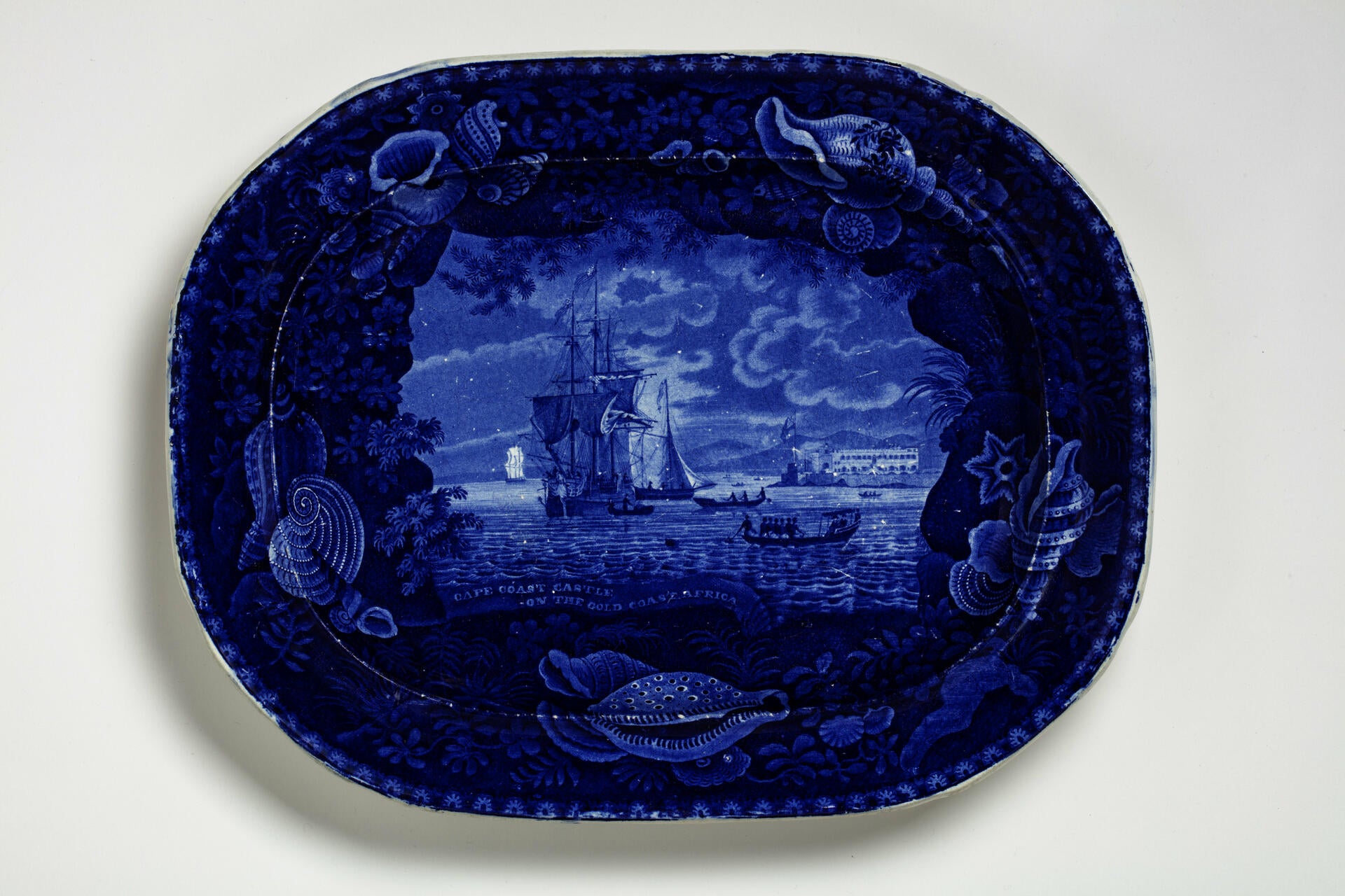 blue and white transferware platter depicting a slave ship off near the Cape Coast Castle, with a decorative shell border