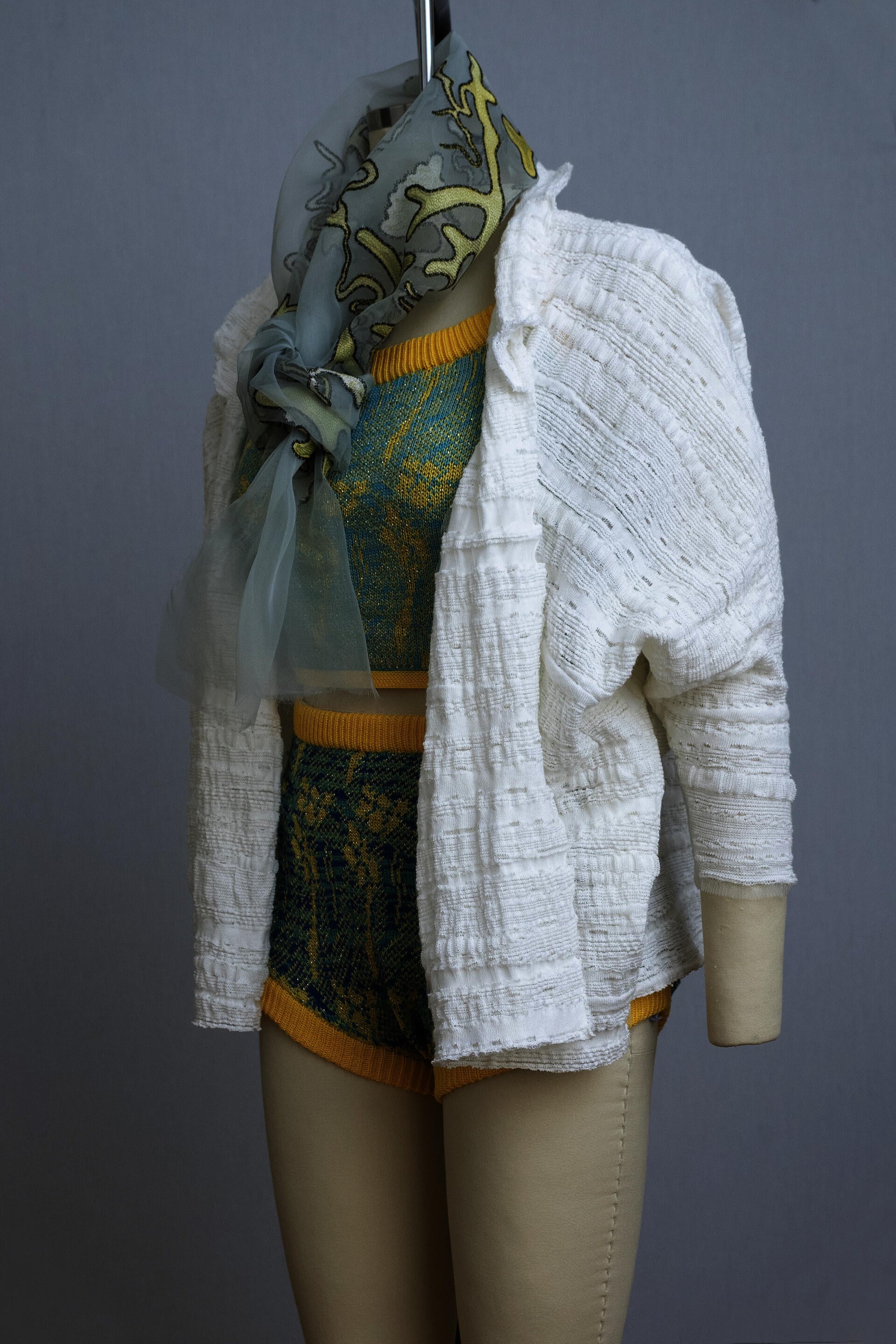 dress form wearing a knit playsuit in blues and yellows, a white knit sweater, and a sheer painted scarf