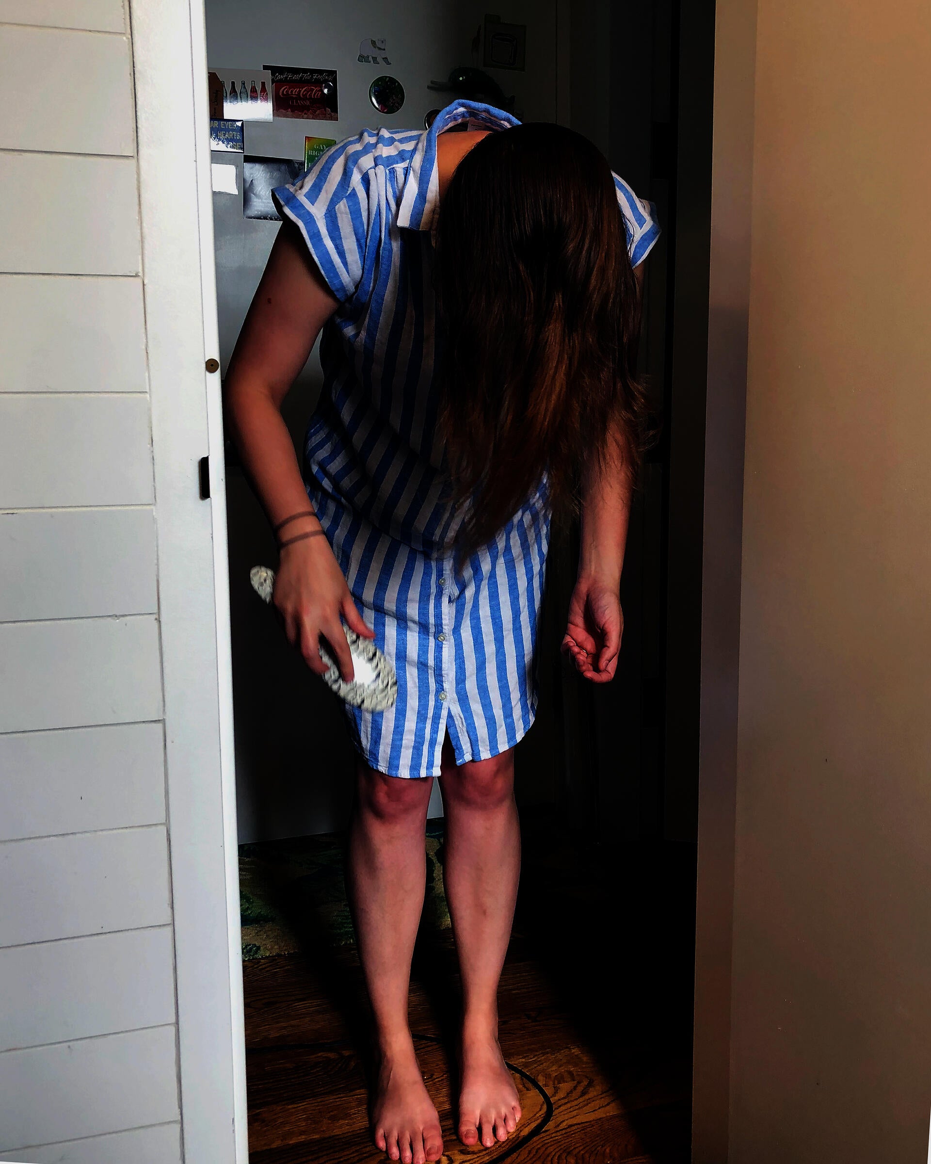 A photograph of a white feminine person in a doorframe. They are barefoot, wearing a blue and white striped shirt-dress and holding a hairbrush, having just pulled it through their long brown hair.