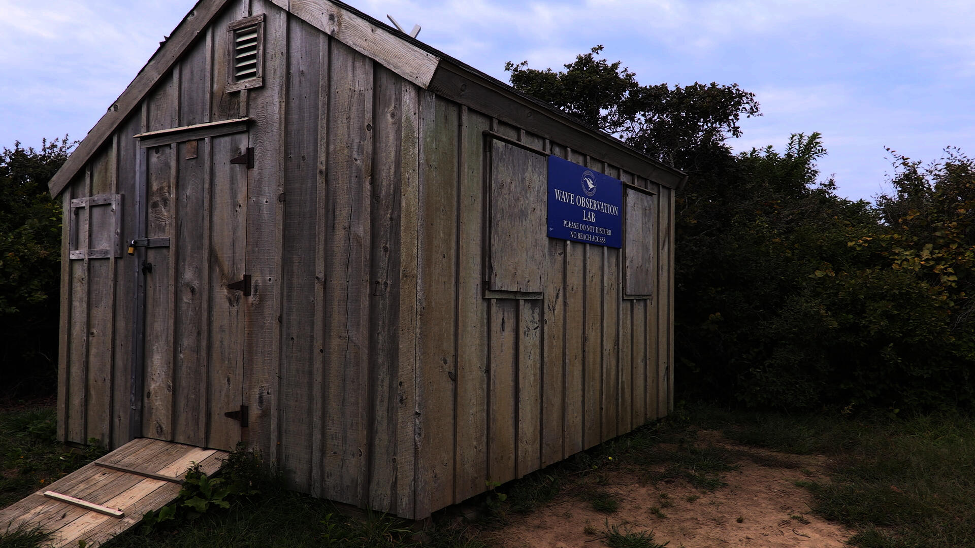 A wooden shed with a blue sign saying Cape Cod Wave Laboratory on a dirt cliff.