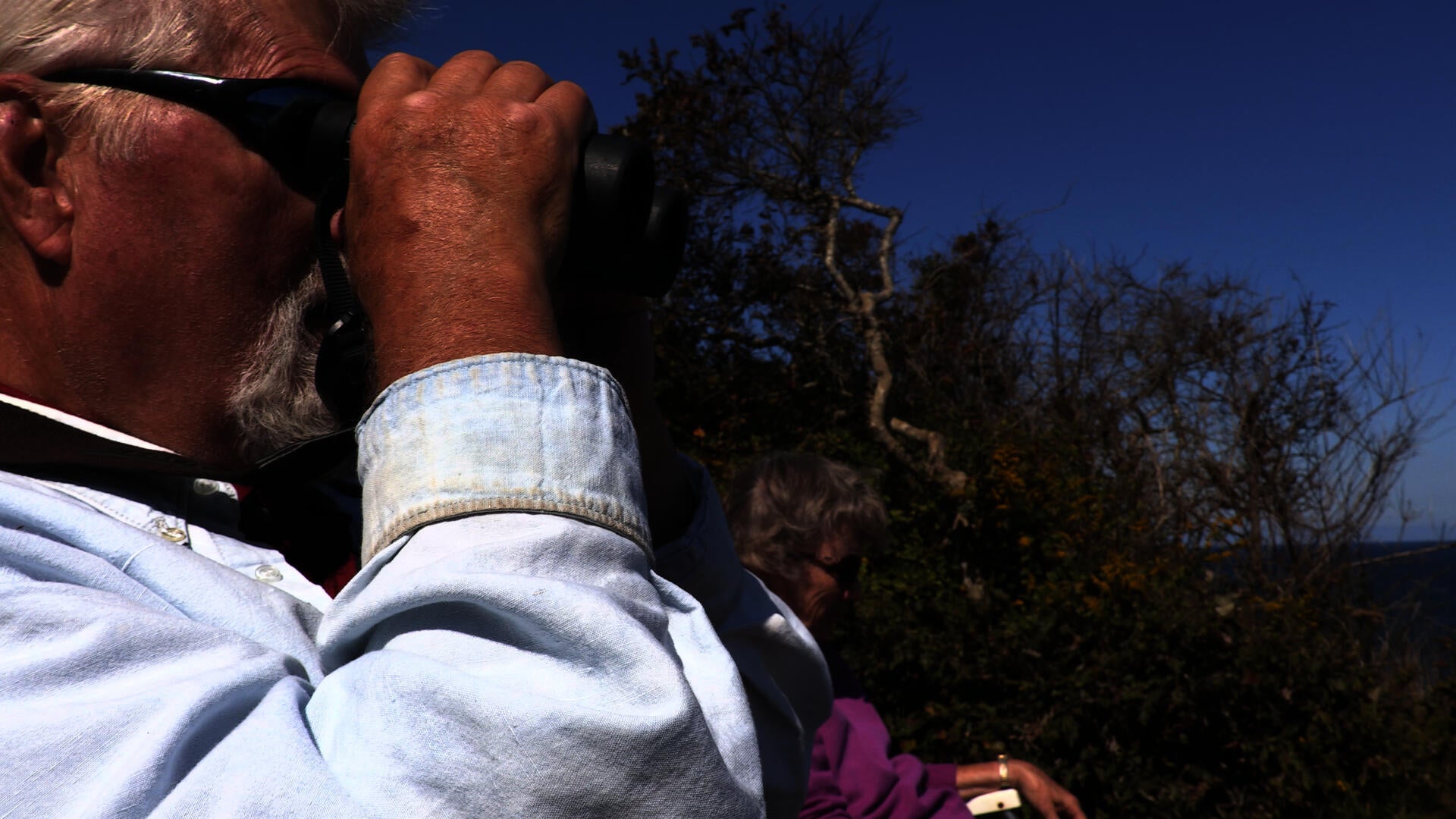 Man sits with his wife and looks through binoculars at the sea.