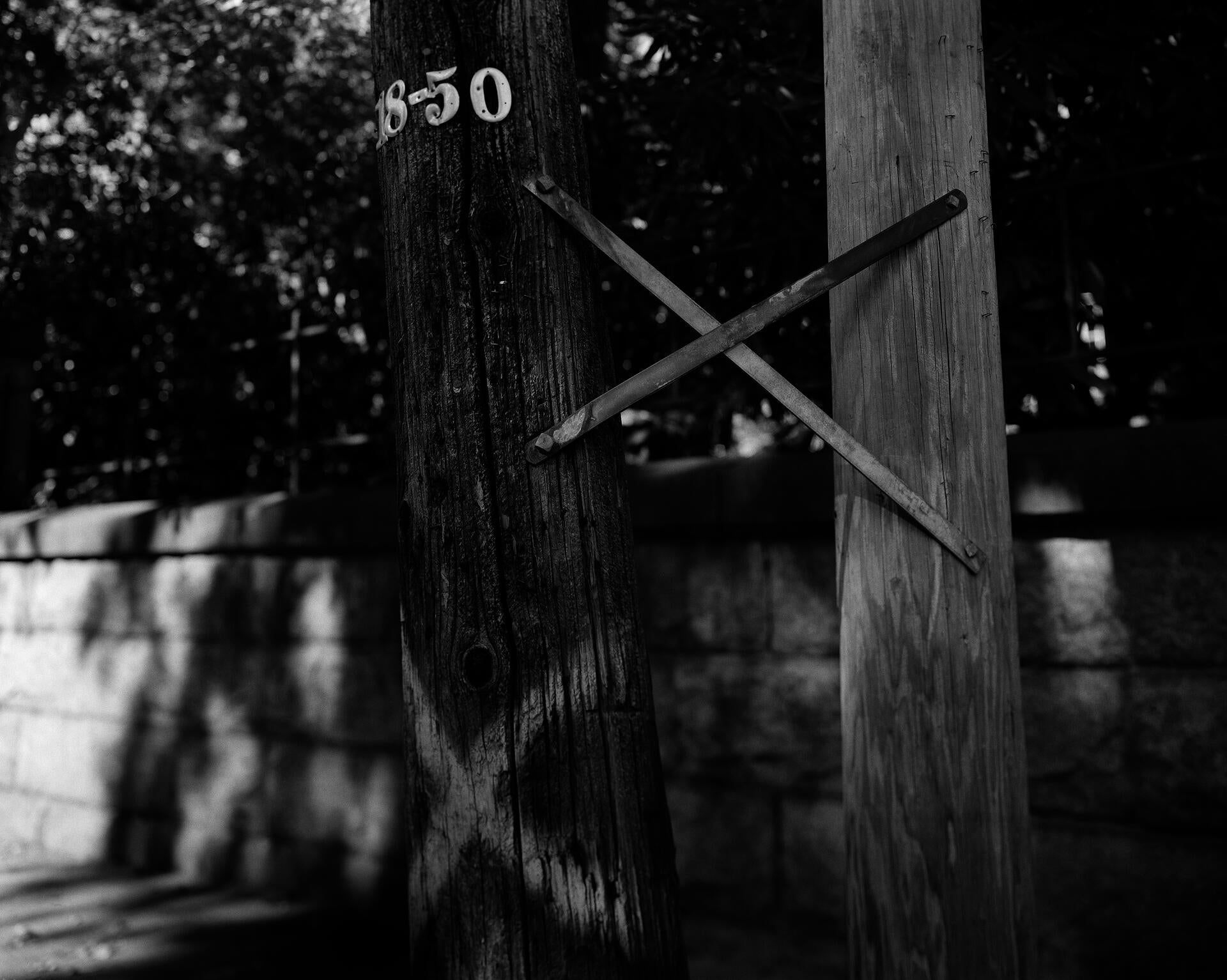 A black and white photograph of two electrical poles bound together with a metal x; one with a painted white x on the lower portion.