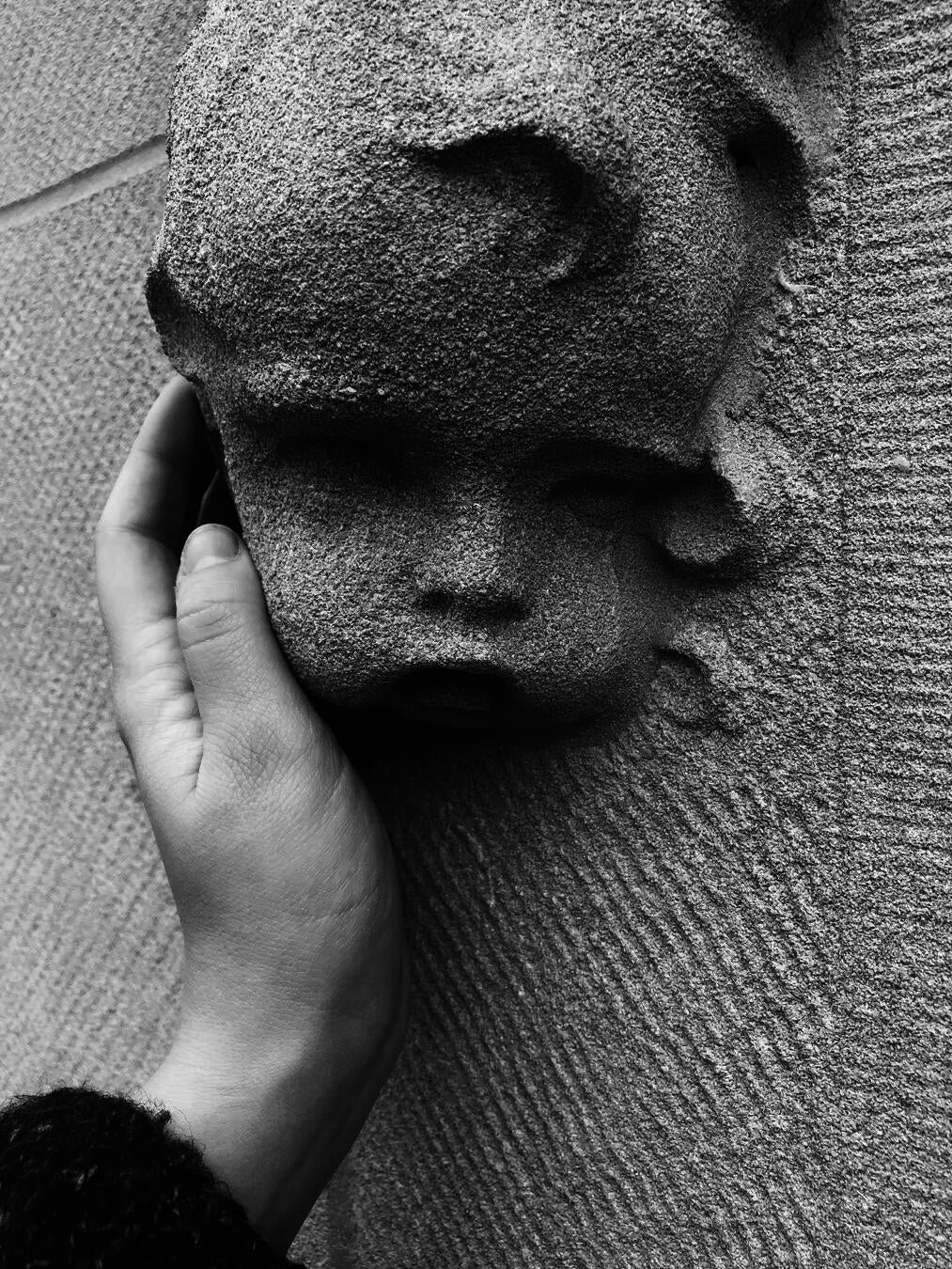 A black and white photograph of a white person's hand reaching out to touch a cherubic face made of stone.