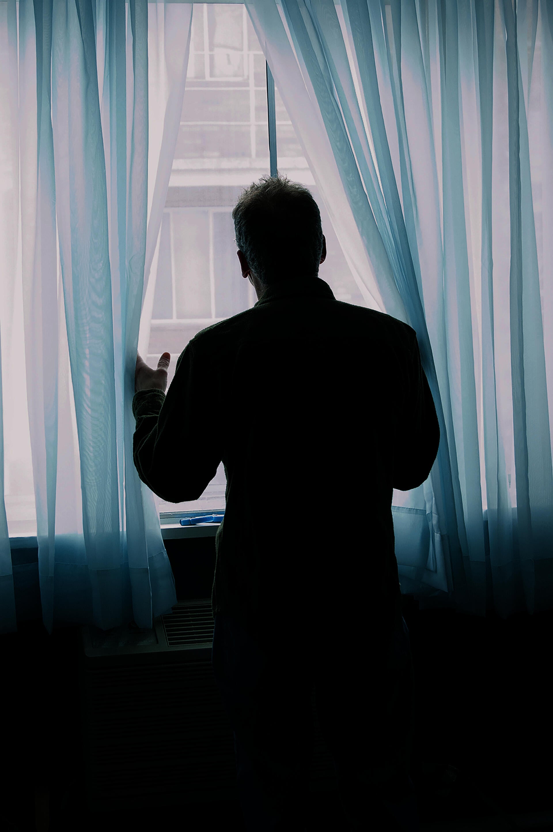 A color photograph of a person looking through translucent blue curtains, seen from behind.