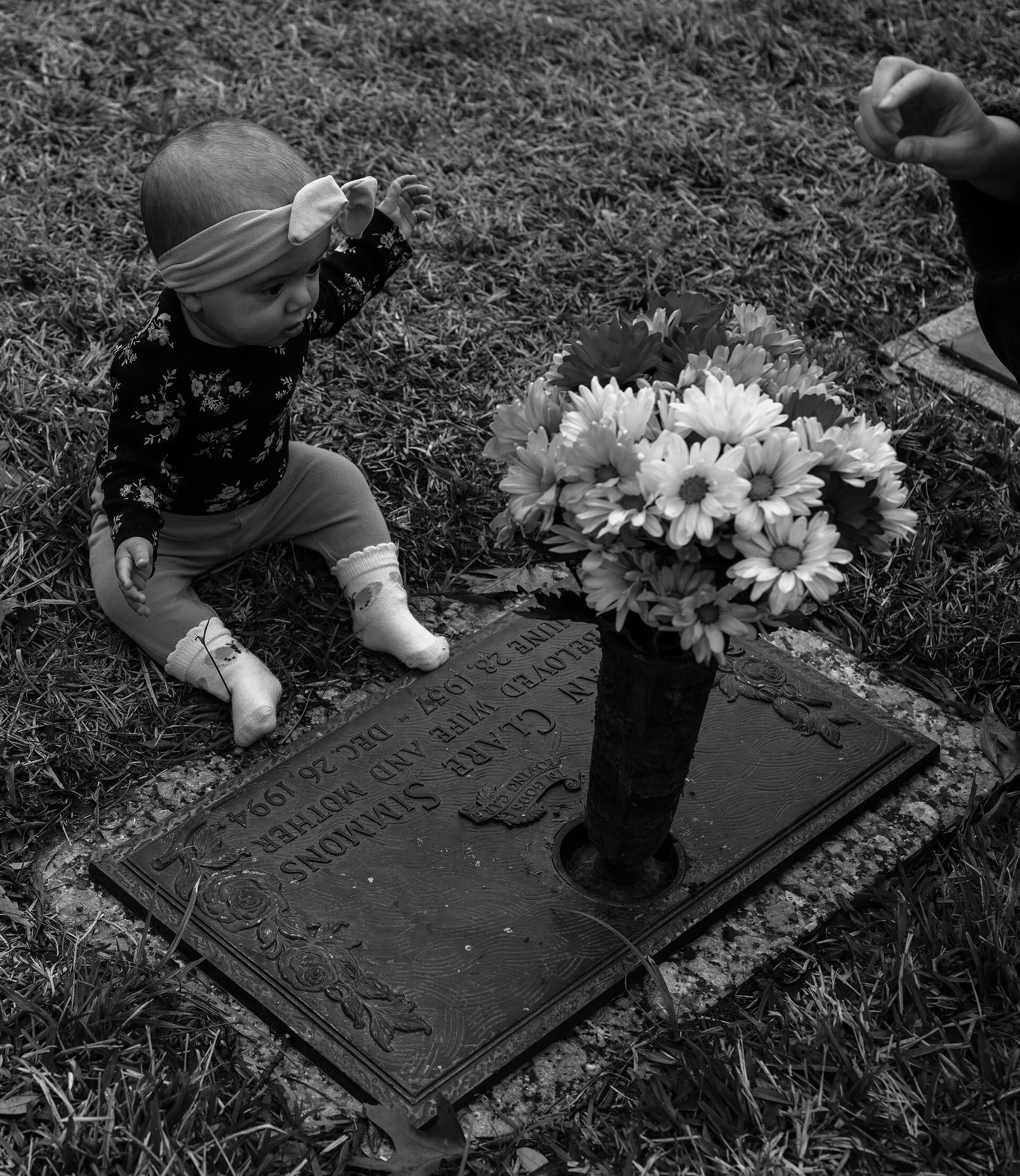 A black and white photograph of a white 6-month-old baby wearing a floral top and a bow, sitting behind a metal headstone. The headstone has a vase on top with multicolored gerbera daisies in it. A white hand is coming into the frame on the right hand side, only visible up to the wrist.
