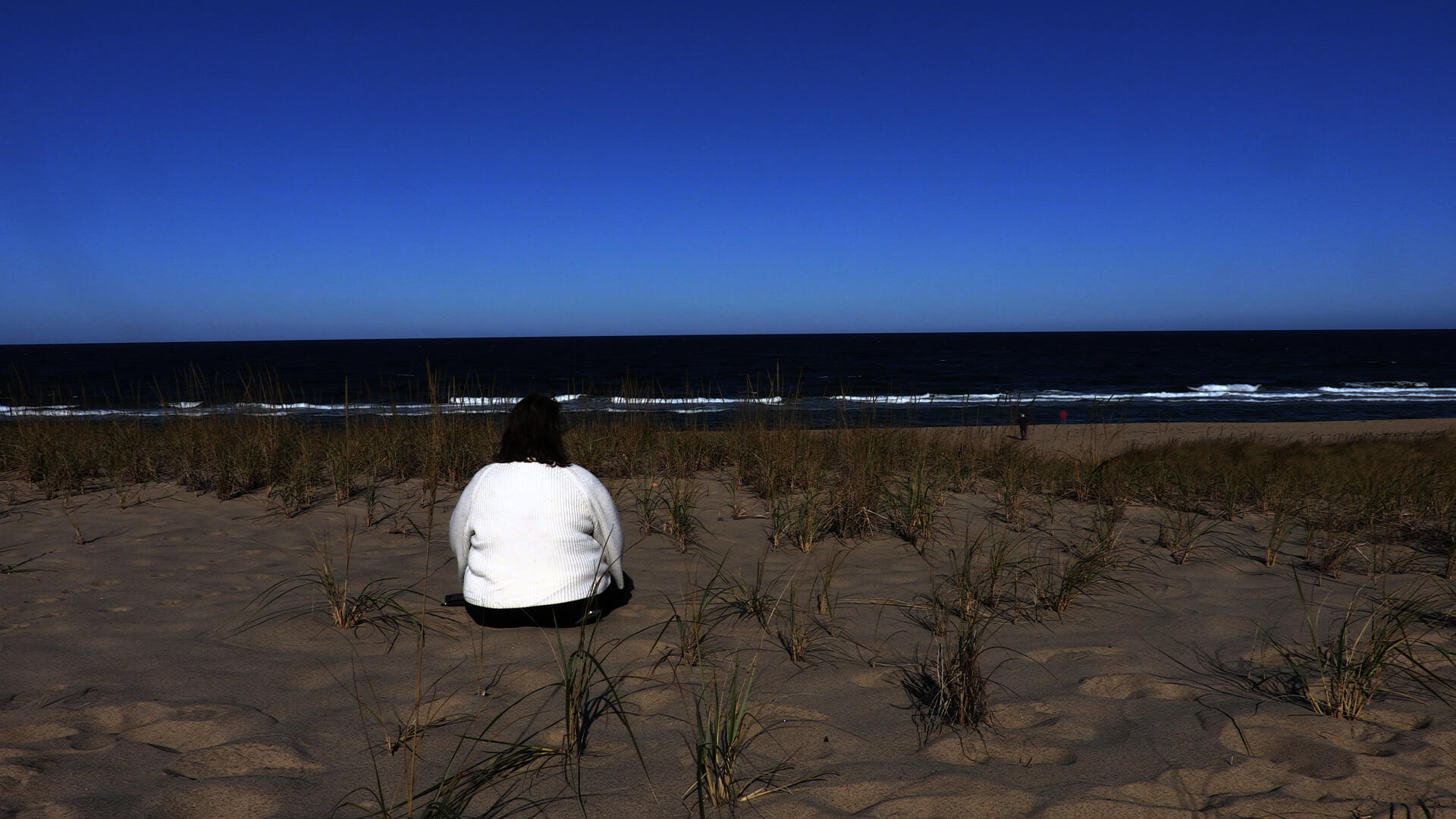 A middle aged woman sitting on a sandy beach with her back to the camera on a sunny day. She appears to be looking at the ocean in front of her.