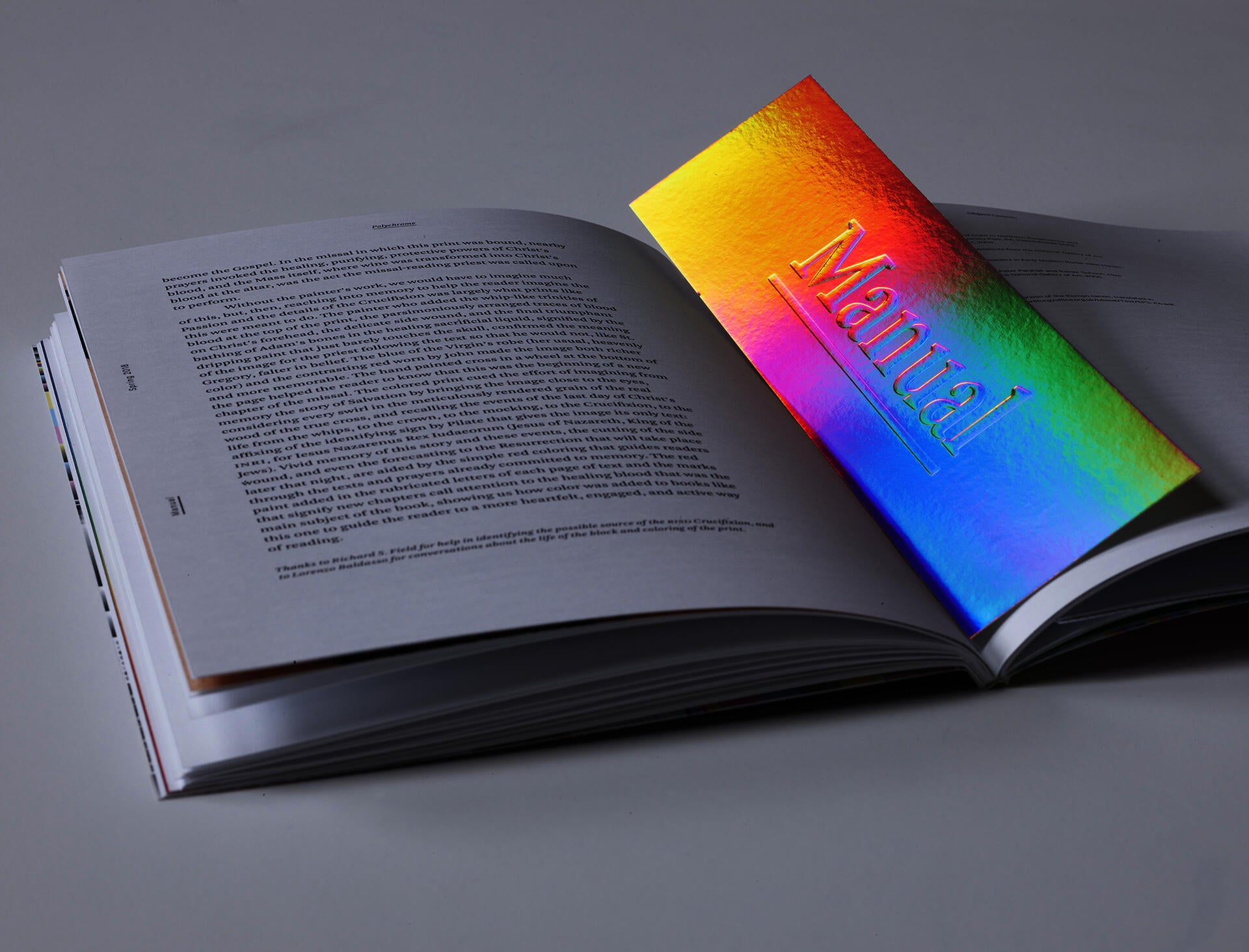 A publication lies open on a white surface. Resting in the middle of the page is a rainbow-colored bookmark, the word “Manual” stamped onto it.