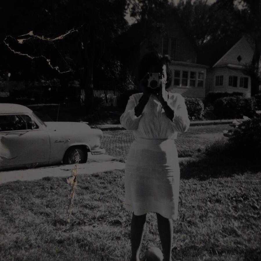Black & white snapshot of a young Black woman dressed in white taking a photograph