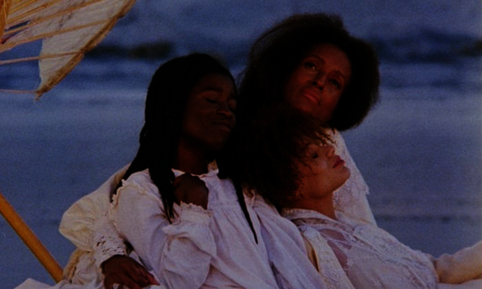 Film still from Daughters of the Dust; three Black women sit dressed in white with the ocean behind them