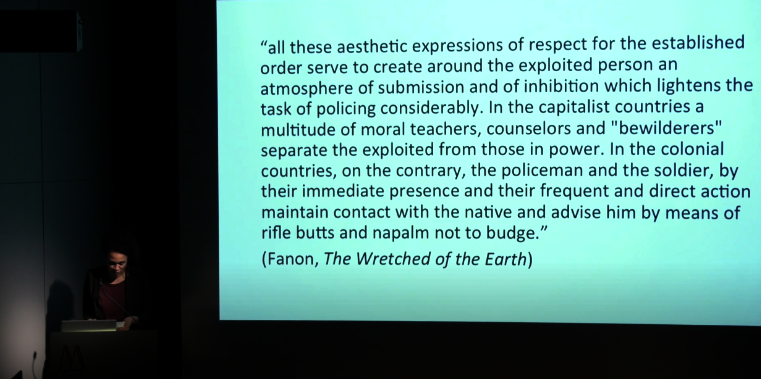 Vanessa Eileen Thompson speaking at the Museum MMK, with a quote by Fanon projected behind