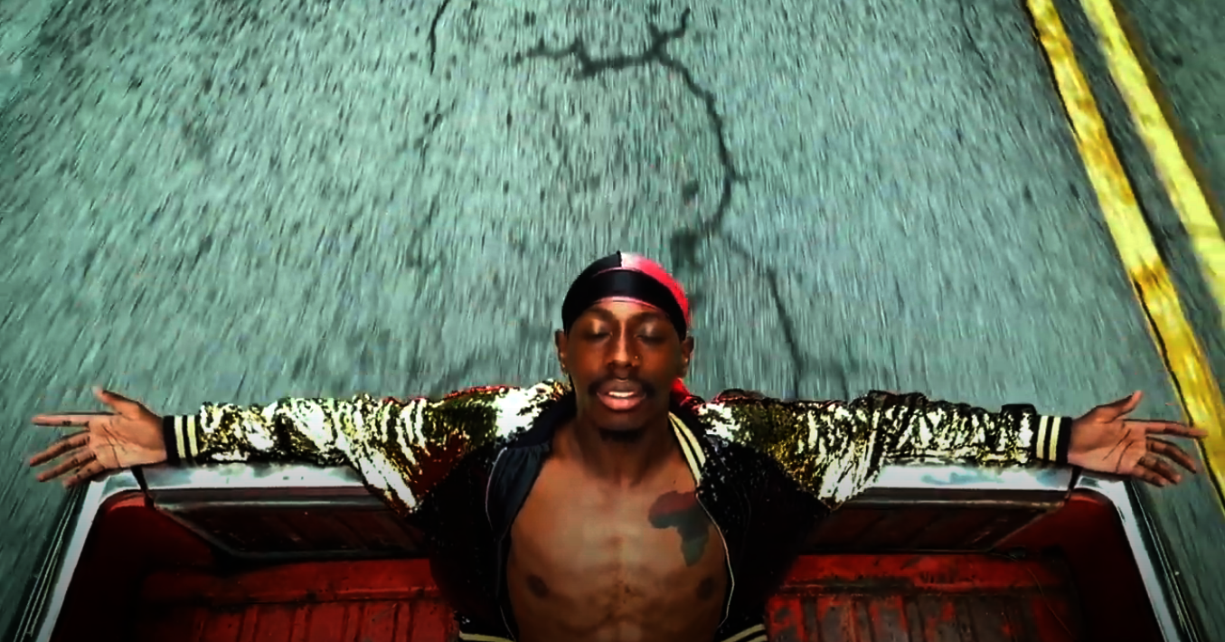 A Black man sits in the back of a truck with his arms outspread and eyes closed, wearing a sequined gold jacket that parts to reveal a tattoo of the African continent on his chest