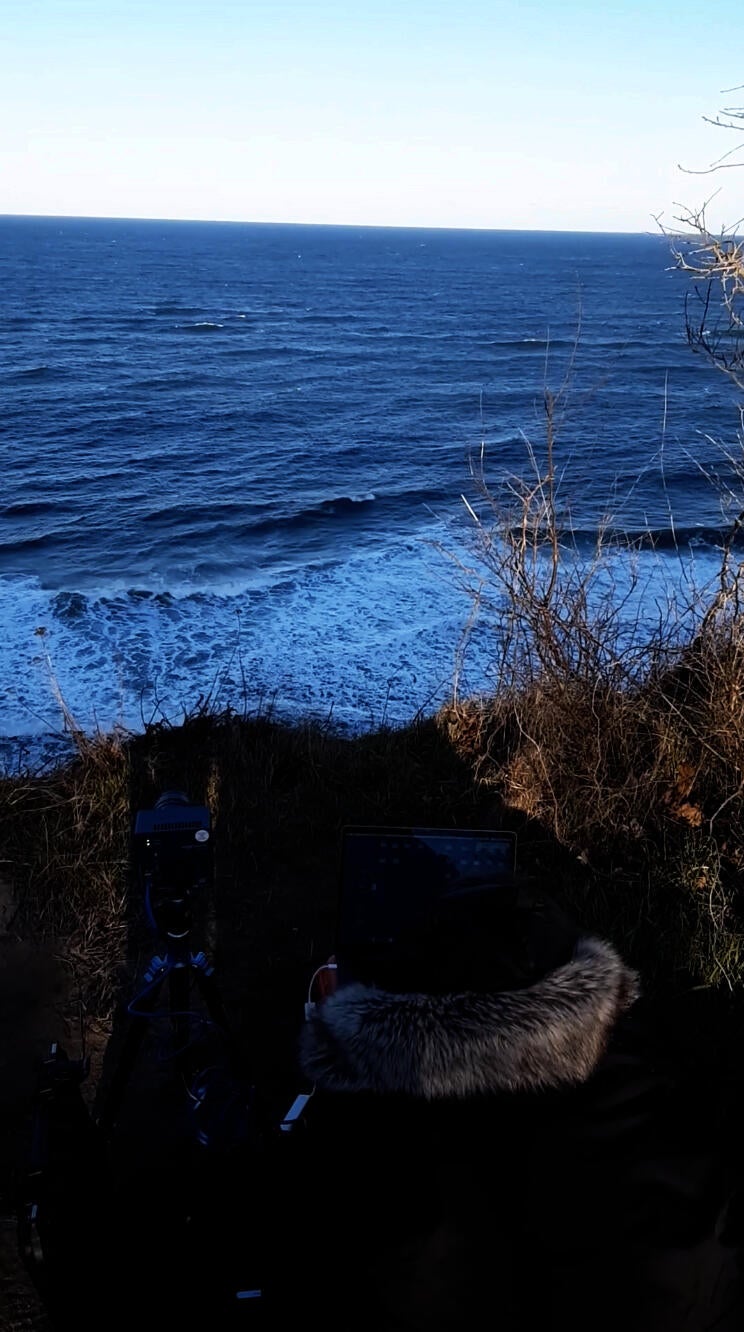 Person in heavy coat works with an Edgertronic Camera and laptop high above the sea.