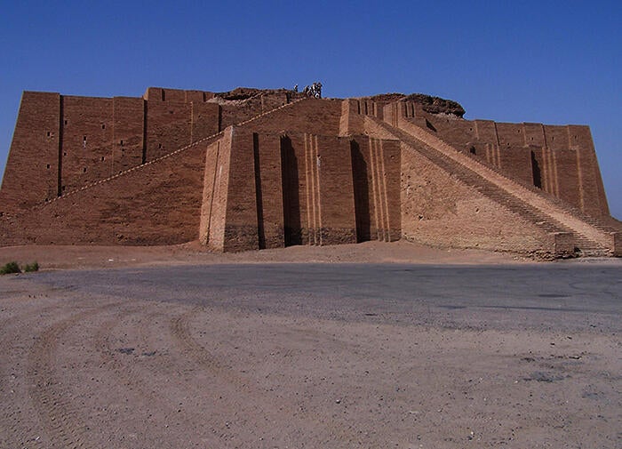 color photograph of the Ziggurat of Ur. Lower half of the photo is beige sand; the upper half is the remains of the Ziggurat and blue sky. The ziggurat a large beige stone rectangular structure with long sloping staircases.