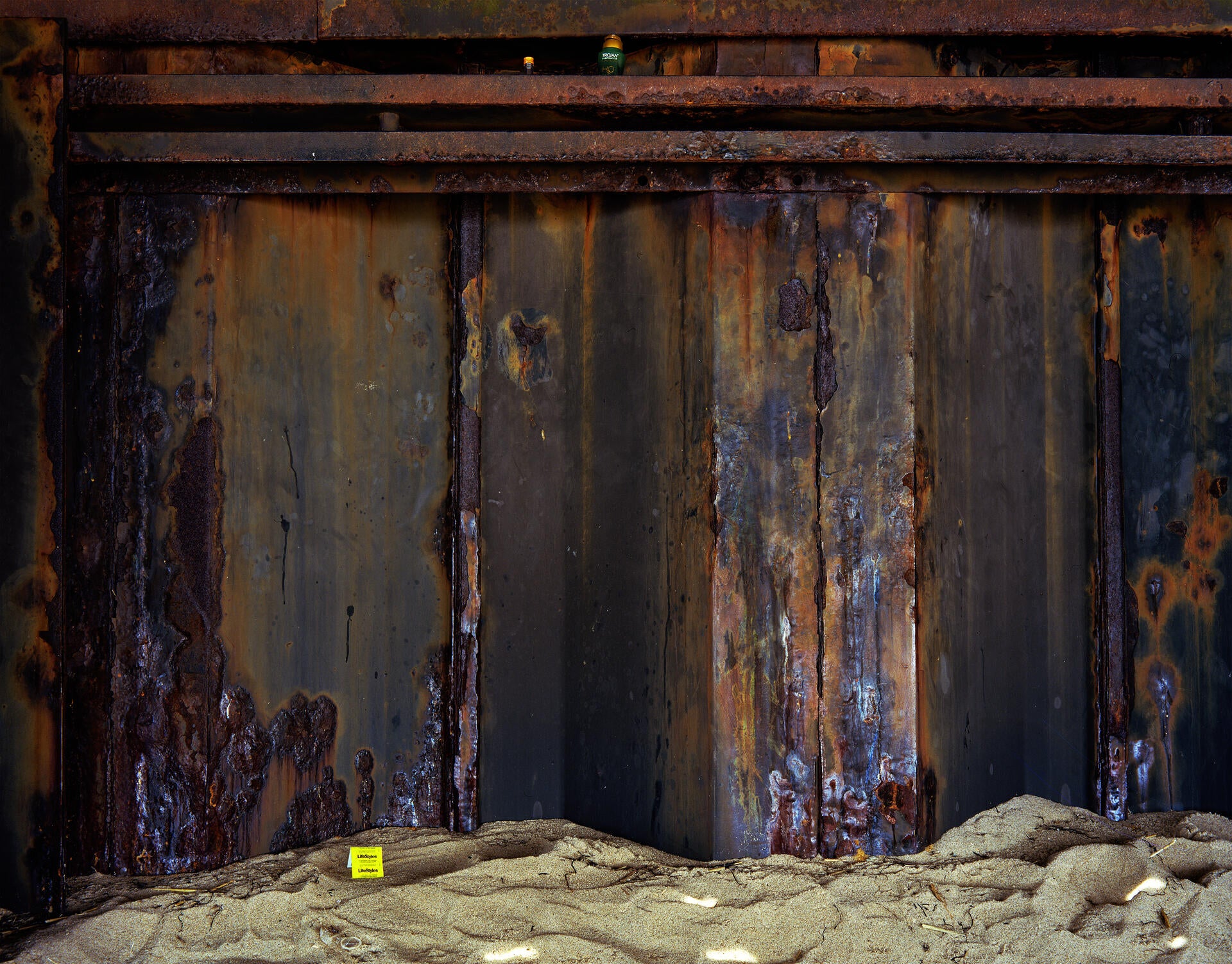 a landscape photograph showing a salt rusted wall with a magnum condom wrapper in the sand. 