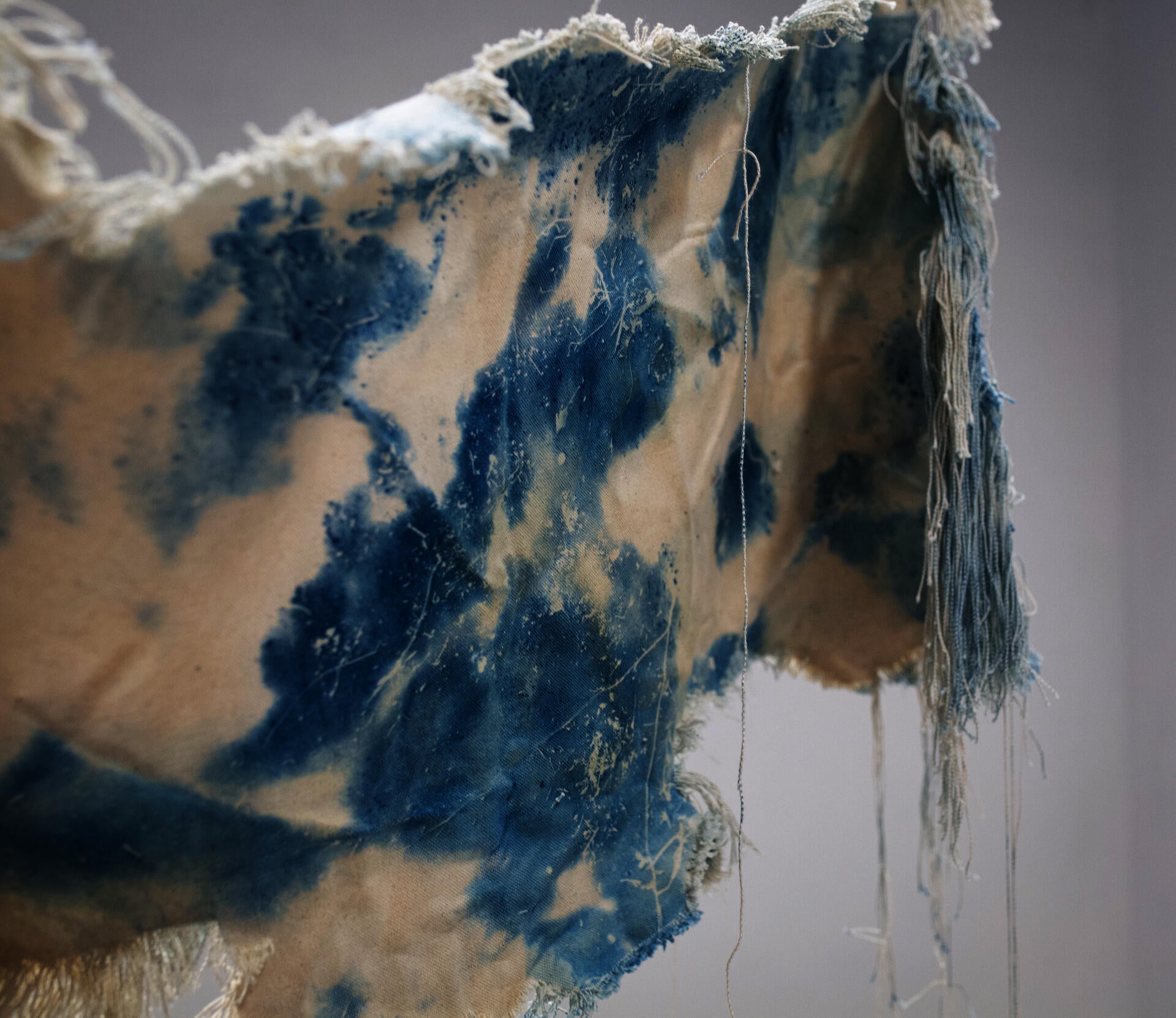 A beige canvas, dyed with spots of cyanotype is twisted and suspended in the air. The edges of the canvas have been unraveled and hang downward.