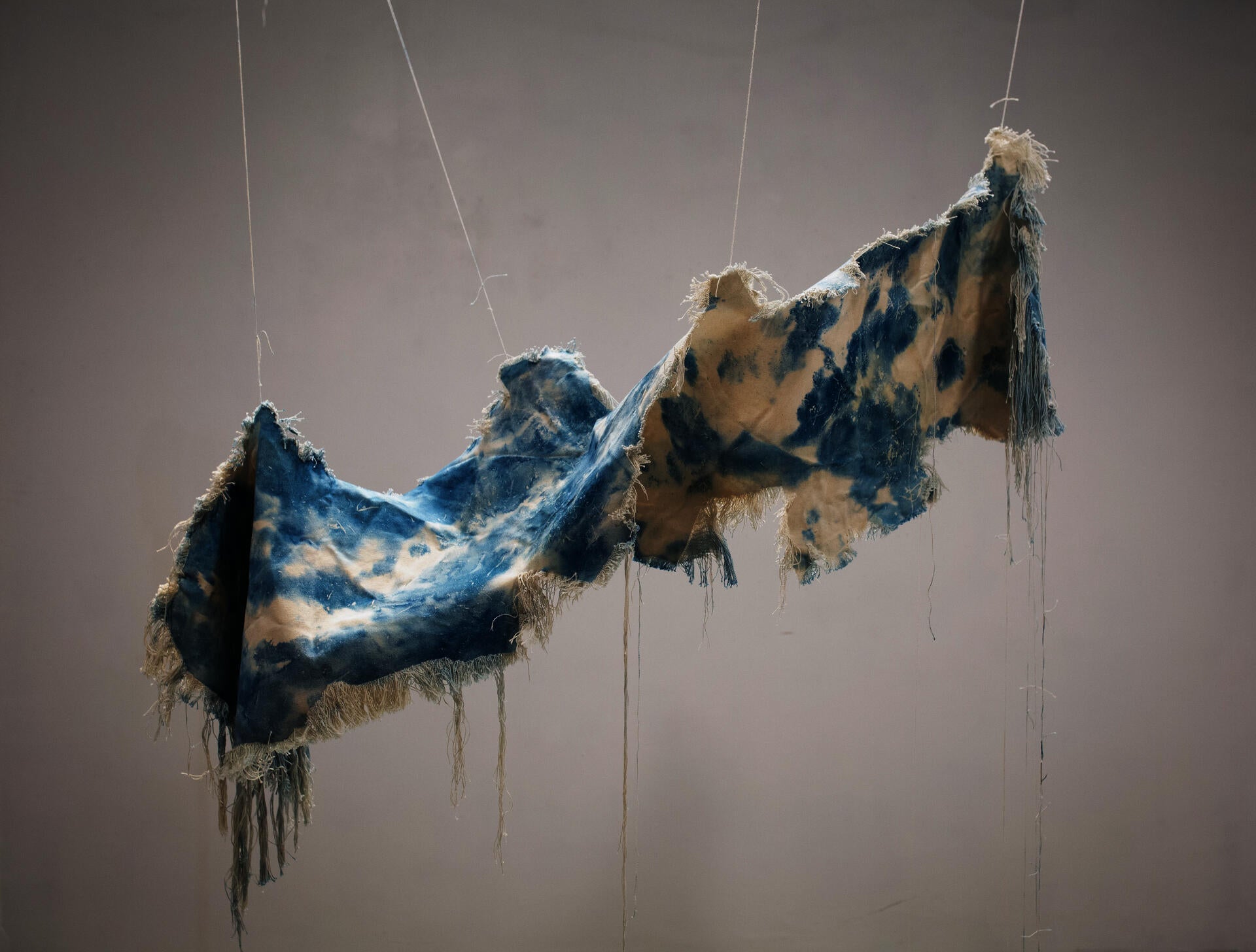 A beige canvas, dyed with spots of cyanotype is twisted and suspended in the air. The edges of the canvas have been unraveled and hand downward.