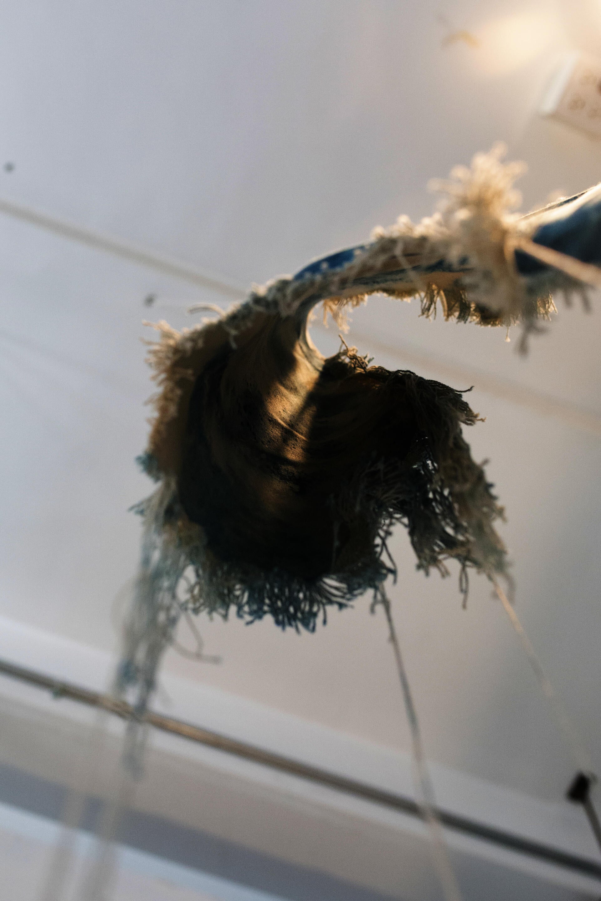A beige canvas, dyed with spots of cyanotype is twisted and suspended in the air. The edges of the canvas have been unraveled and hang downward. 