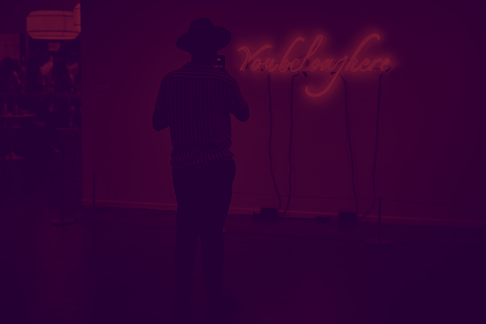 Standing with back to the viewer and in shadow, a person photographs a neon sign. It says, in light blue script, “You belong here.” A purple filter has been applied to the photo.