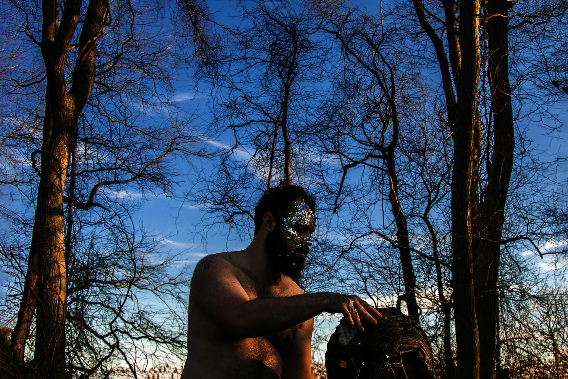 Photograph of artist in forest holding handmade nest made out of clay with plastic  silver confetti on his face. 