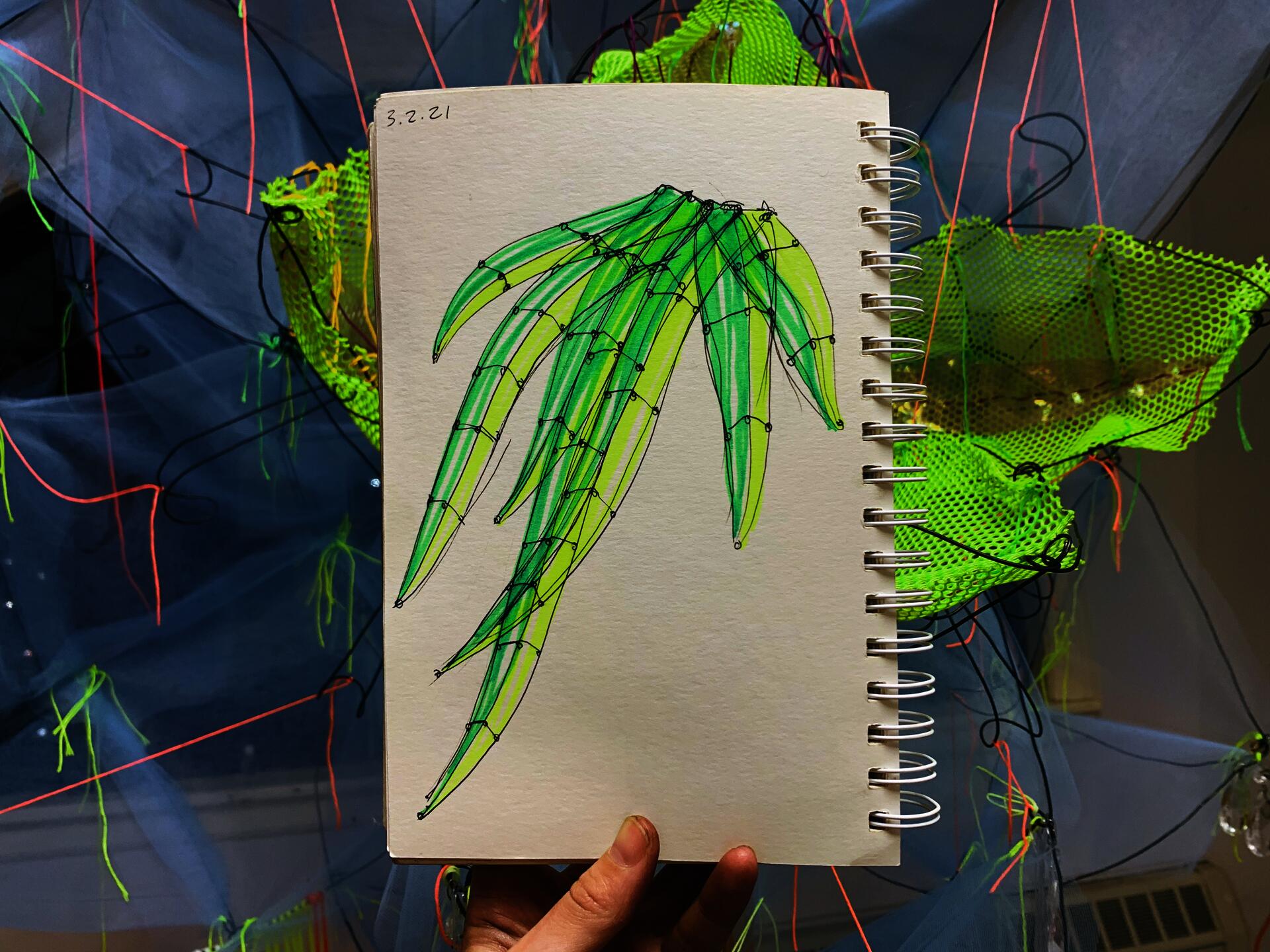 Sketch of how blades of grass can be turned into a wire sculpture. Page held by hand in front of a wire and fabric sculpture of a magnolia. - The Ephemeral Altar & The Floating Grave - Hannah Suzanna