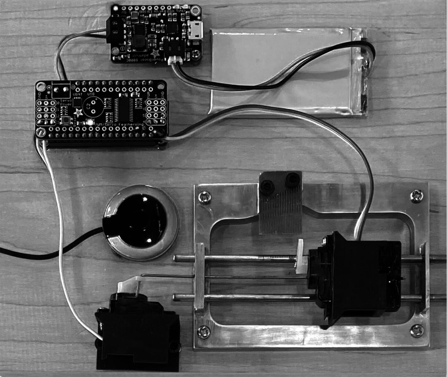Image of a robotic music box. Two small circuit boards, a flat, rectangular battery, a round microphone, and a small motor are laid out around a metal frame which holds the music box tines and a motor on a track.