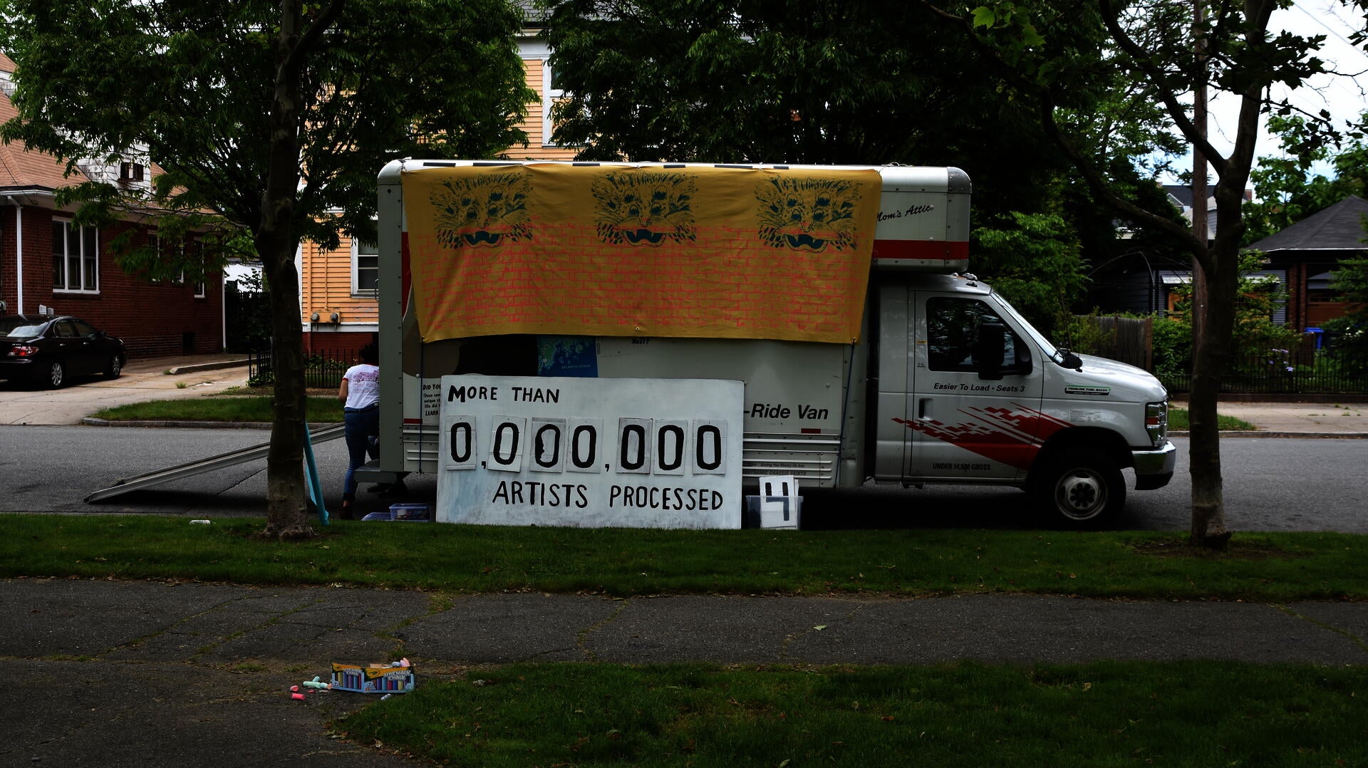 A U-haul van is parked by a park and some houses. A large banner with a brick pattern and three four-eyed lions hangs from the truck, and a big sign that says 'More that 0,000,000 artists processed' with replaceable numbers leans against the truck.