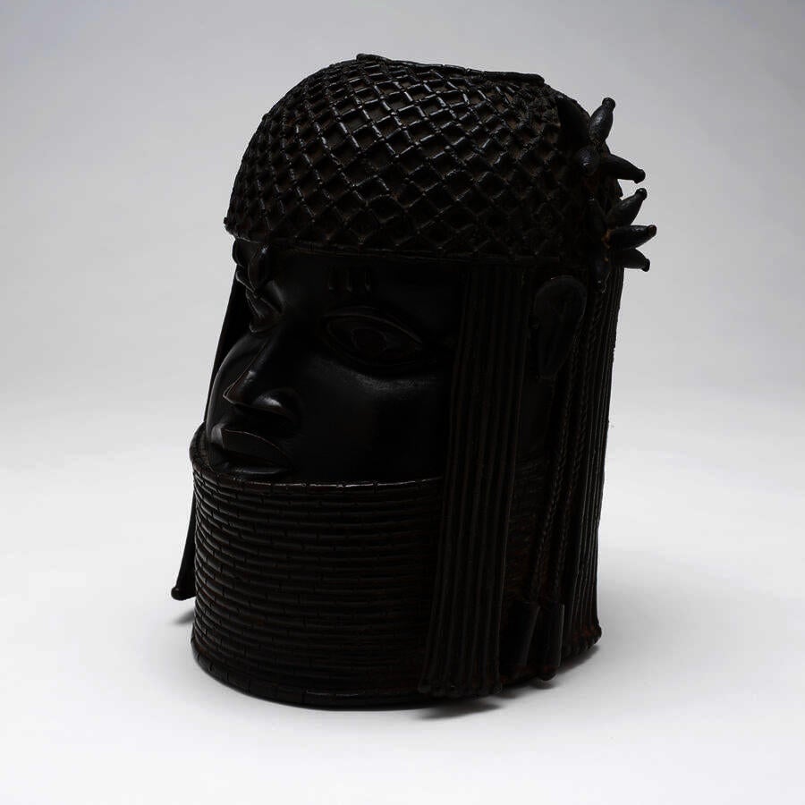 Elegant stylized portrait head. The subject wears an elaborately patterned headcovering and a tall stack of necklaces. Color is dark brown.
