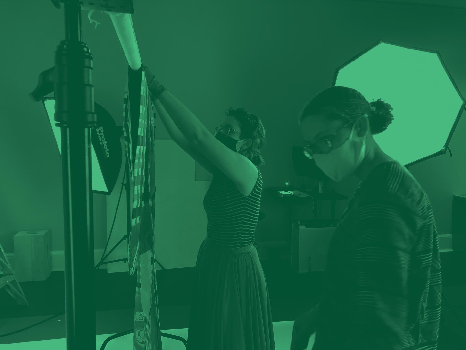 Two masked people set up photography for a textile. A large lamp shines behind them. A green filter has been applied to the photo.