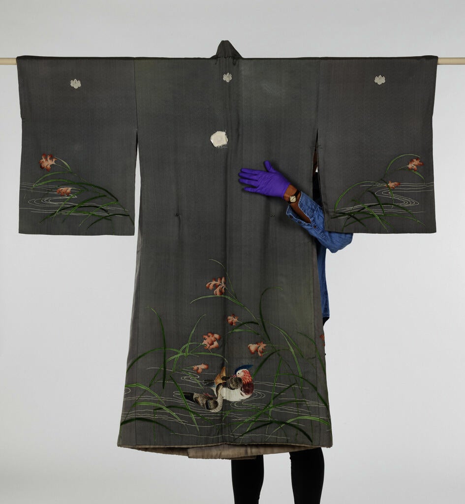 An embroidered gray Asian robe hangs from a rod. A museum worker stands behind it, looping one purple-gloved hand to the front. The result is something like an embrace.