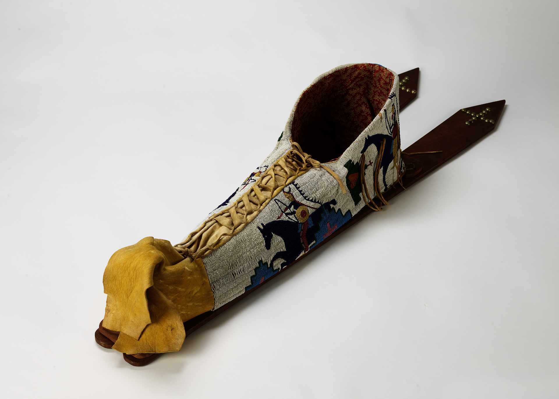 Beaded baby carrier lying flat, shown from above. Back boards angle to the right. Top is open, front center laces up with leather ties, bottom is loosely folded.