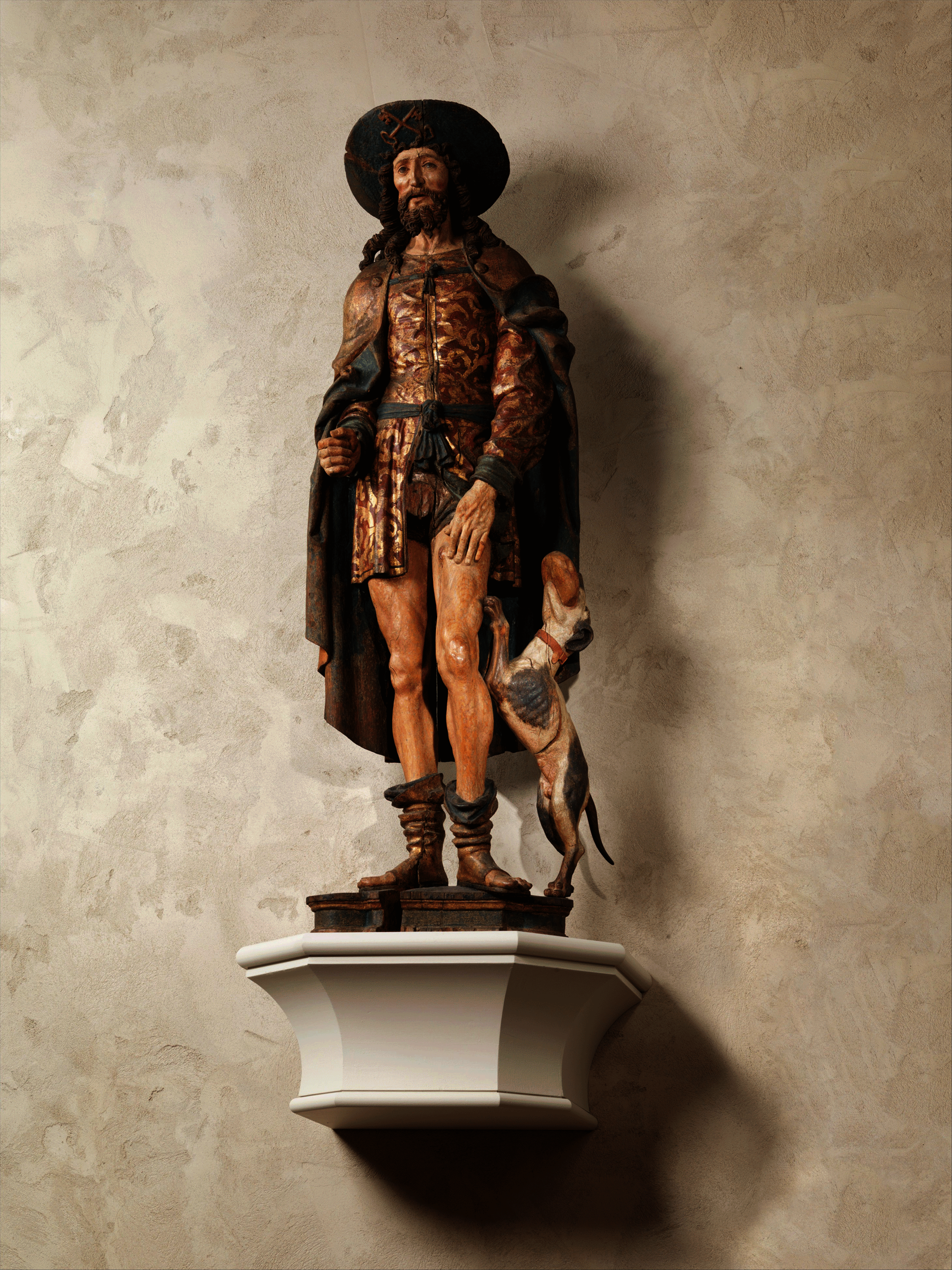 Painted sculpture of a bearded man in an elaborate short tunic and cloak. His left hand rests on his bare thigh. A dog stands on his back legs beside him.