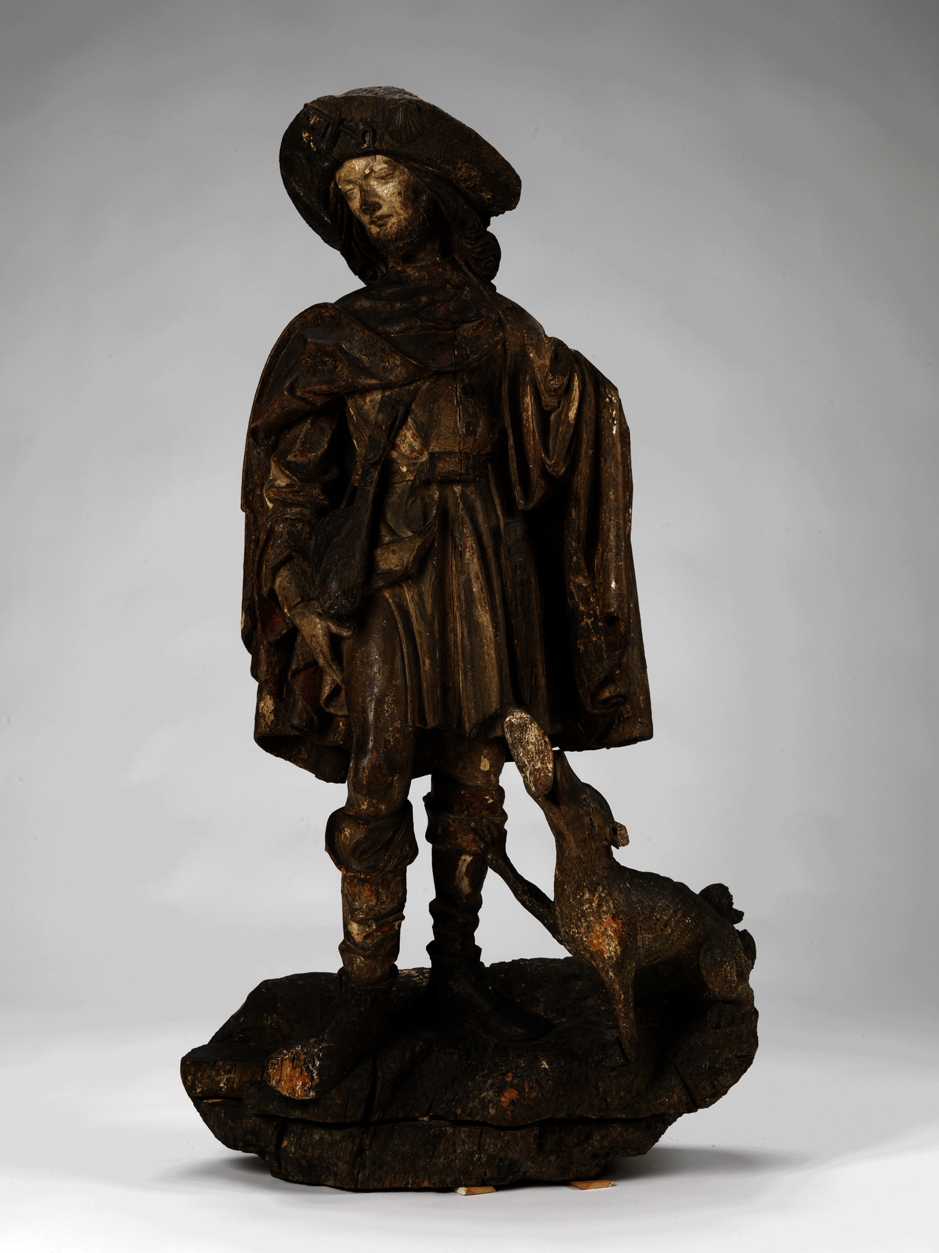 Dark-brown sculpture of a standing man wearing a cape, hat, and boots. He looks to the side, hand on his thigh. A dog at his feet looks up at him.