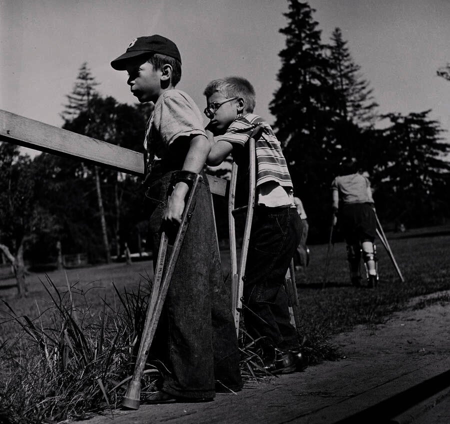 Two light-skinned boys stand outside in profile, supported by crutches, watching something out of frame. Behind them, another person in leg braces walks away using crutches. 