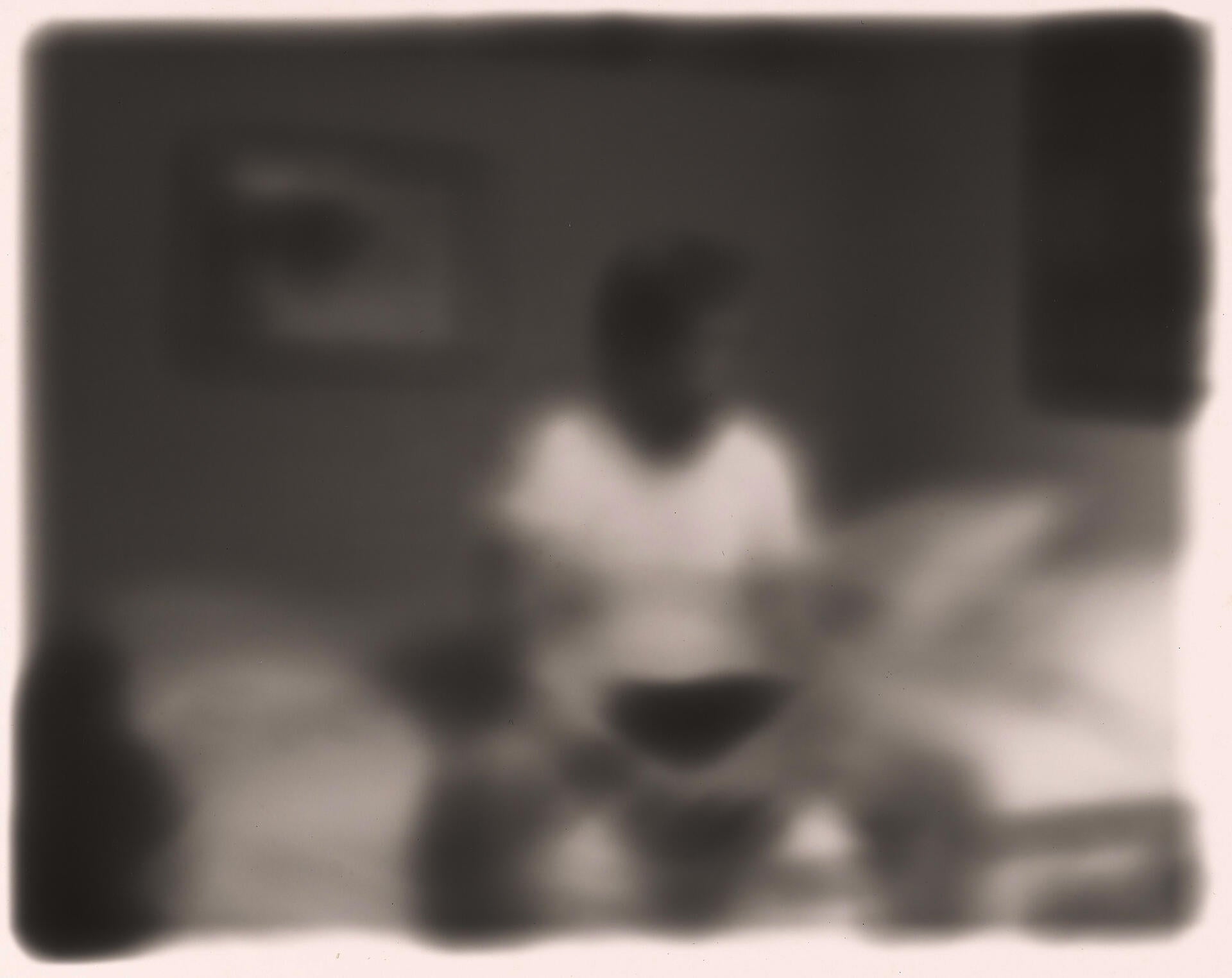 Dreamy, out-of-focus black-and-white image of a figure sitting on a bed, hands clasped between their knees.