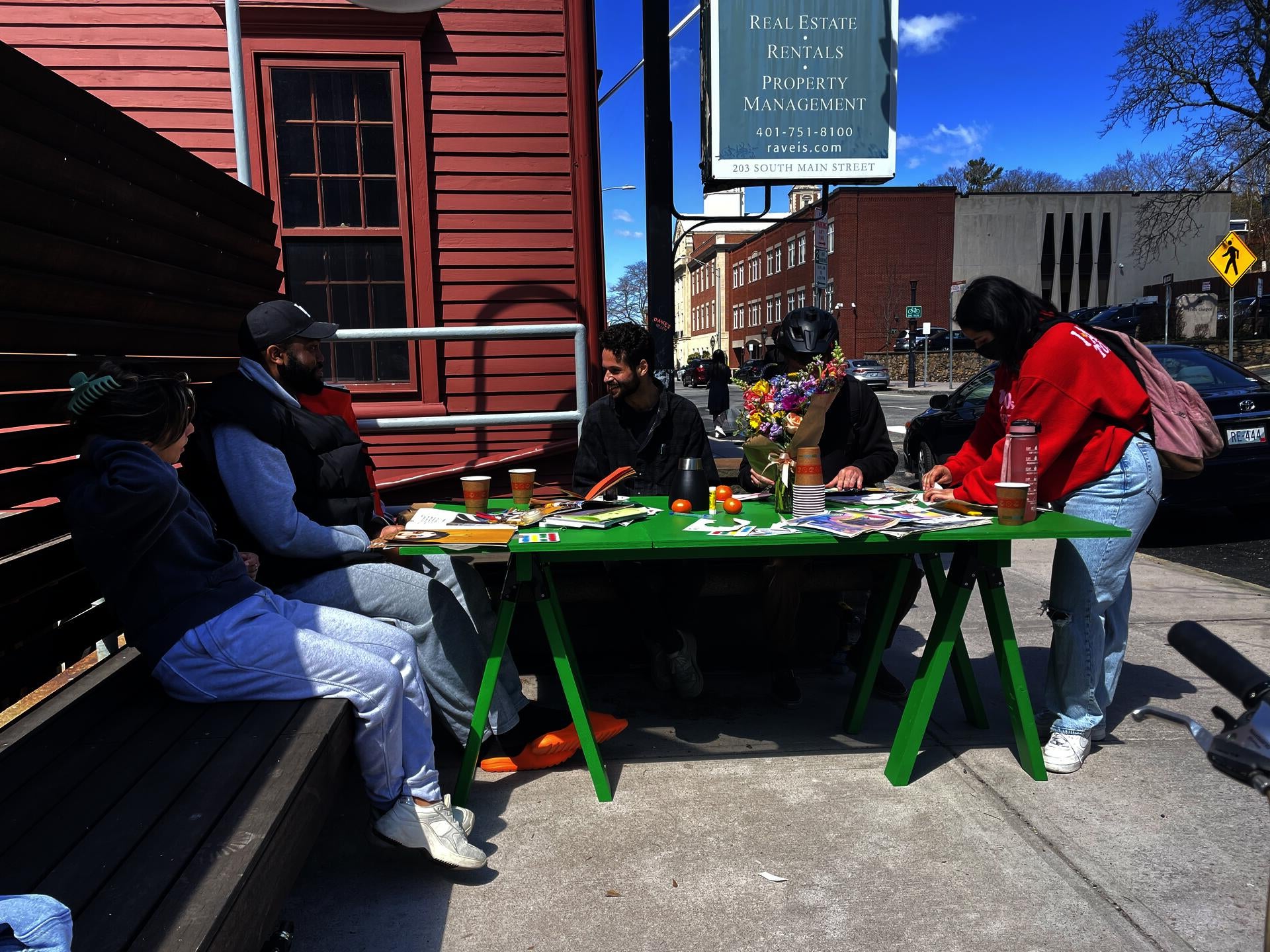 Five students gather around a bright green table that is covered in collage materials, flowers, drinks, and citrus. 