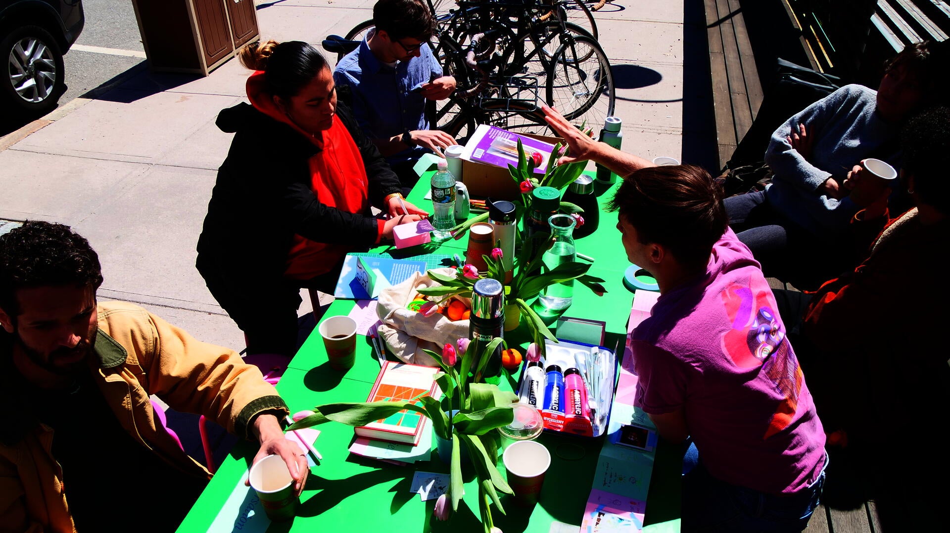 6 students gather around a green table covered in flowers, drinks, paint, and a books of drawings and photographs.  