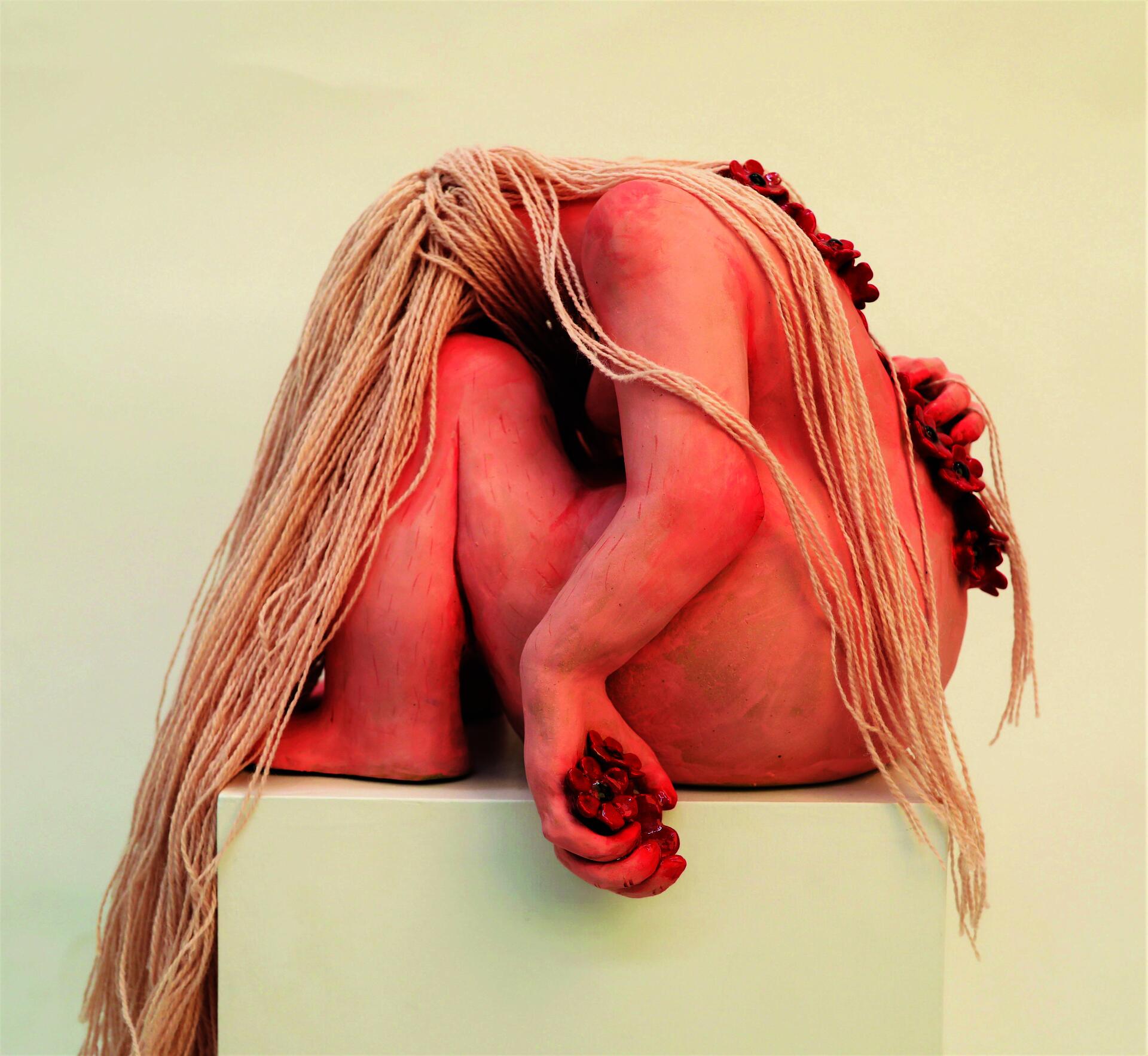 A pink ceramic figure with pink yarn hair rests its head on its knees, looking away.