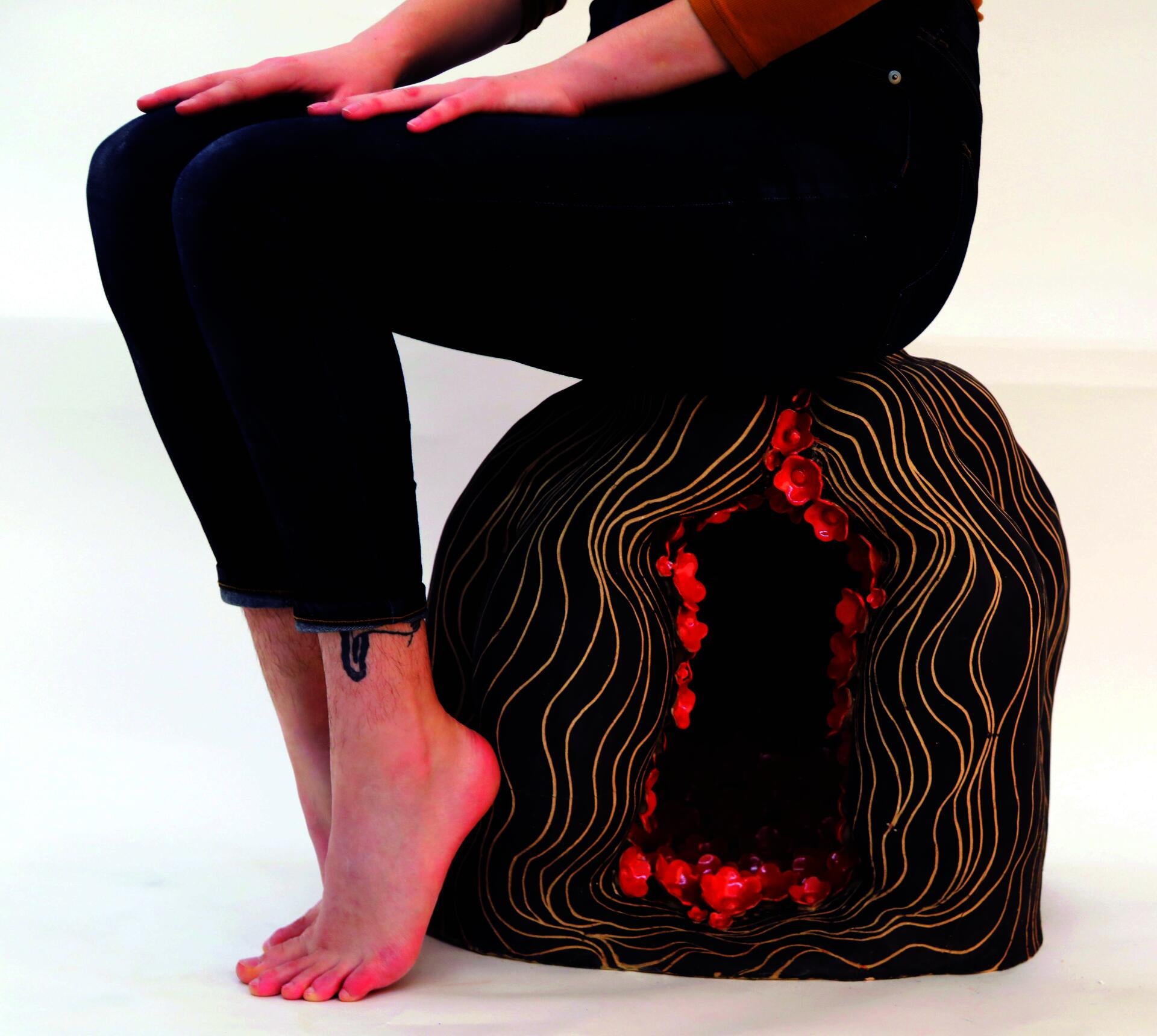 Image of a person from the waist down sitting on a ceramic head with red flowers inside its cavity.