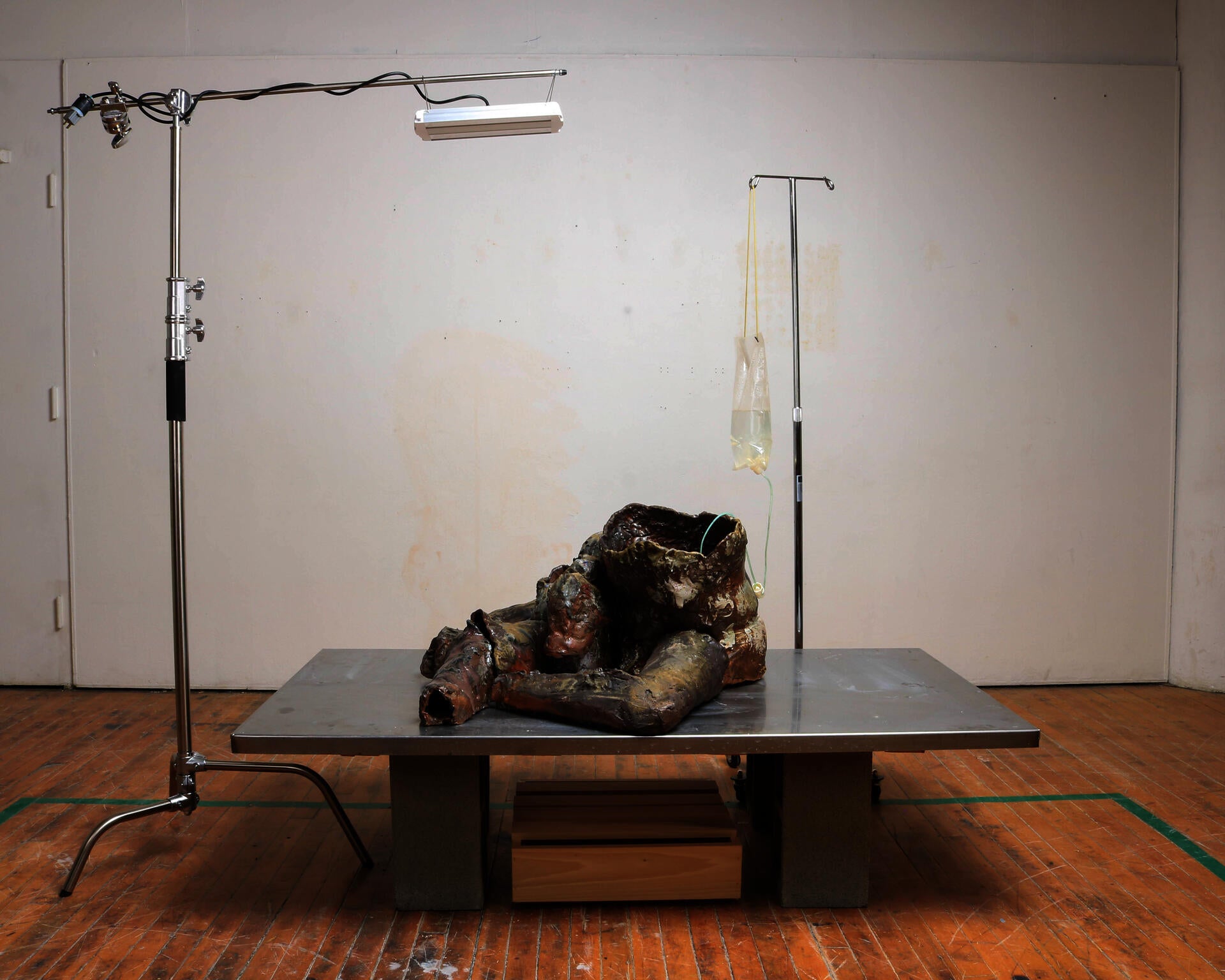 A dark, abject ceramic body with only two abstracted legs sits upon a steel slab with an IV Bag and grow light above.