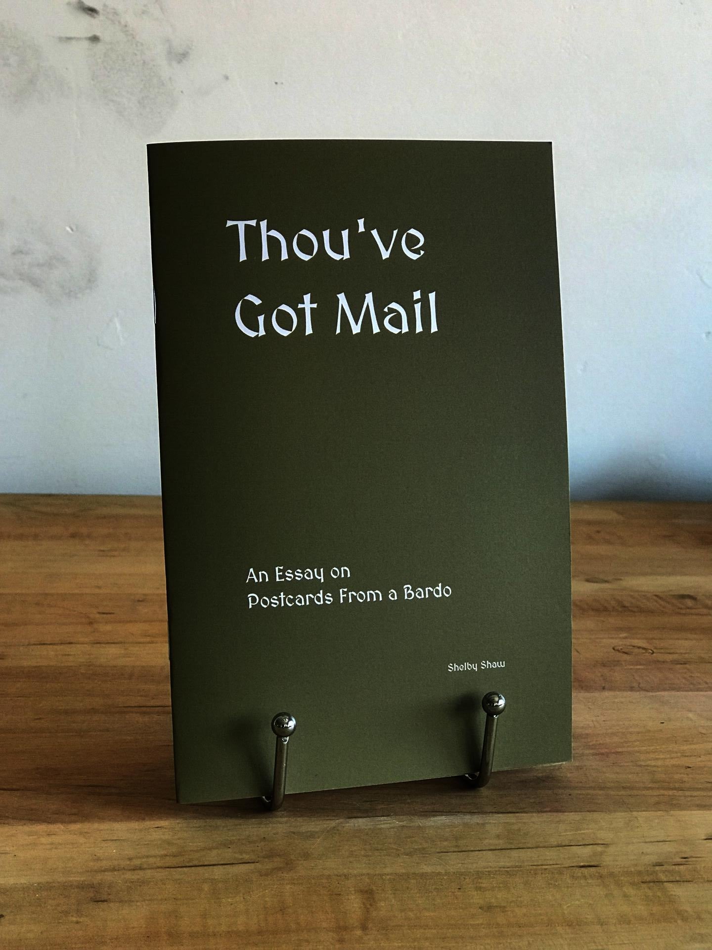 The book 'Thou've Got Mail' which is a khaki green-brown color with white font is displayed on a silver book stand atop a wooden tabletop with a white wall in the background.