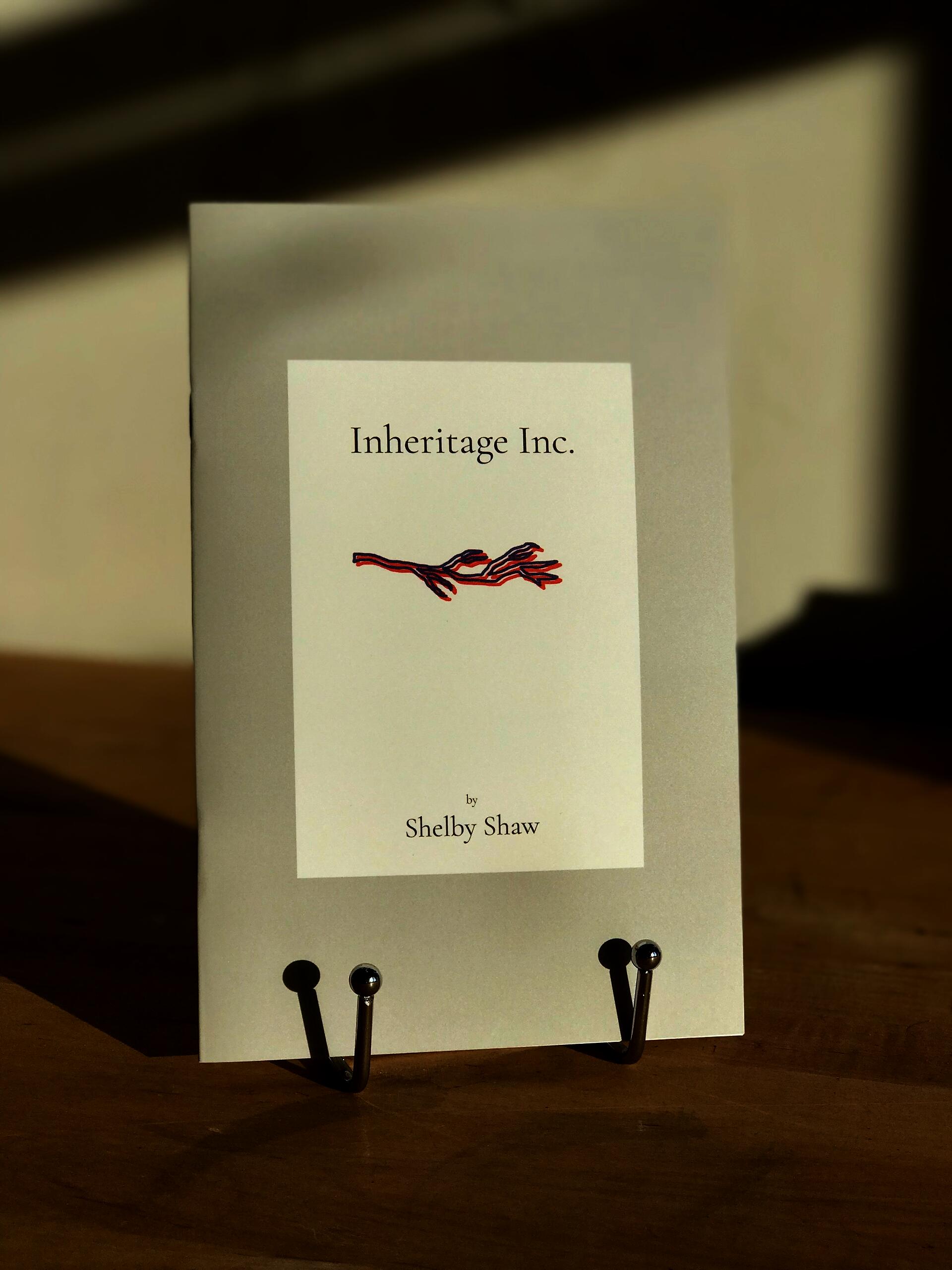 The book 'Inheritage Inc.' which is a grey color with white center rectangle containing the title in black font and a red and blue illustration of a tree branch, is displayed on a silver book stand atop a wooden tabletop with a white wall in the background.