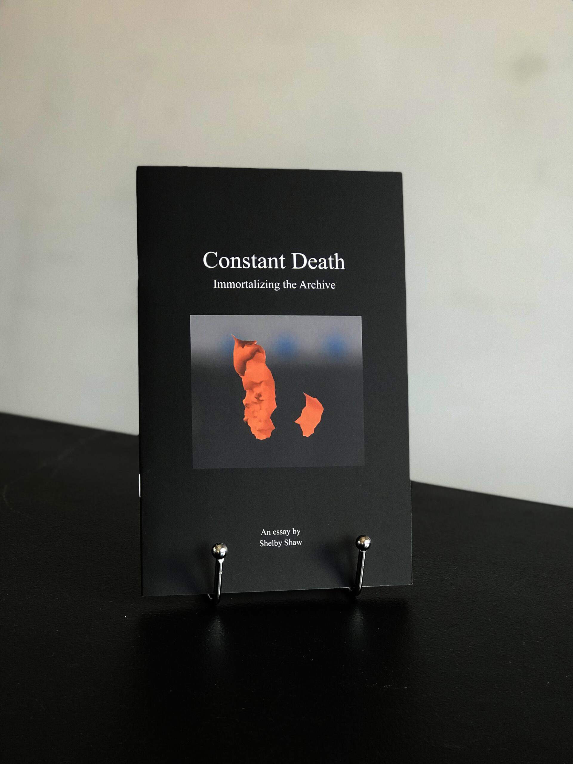 The book 'Constant Death' which is black with white font for the title placed above an inserted image of a strange brown shape on a blue background, is displayed on a silver book stand atop a black tabletop with a white wall in the background.