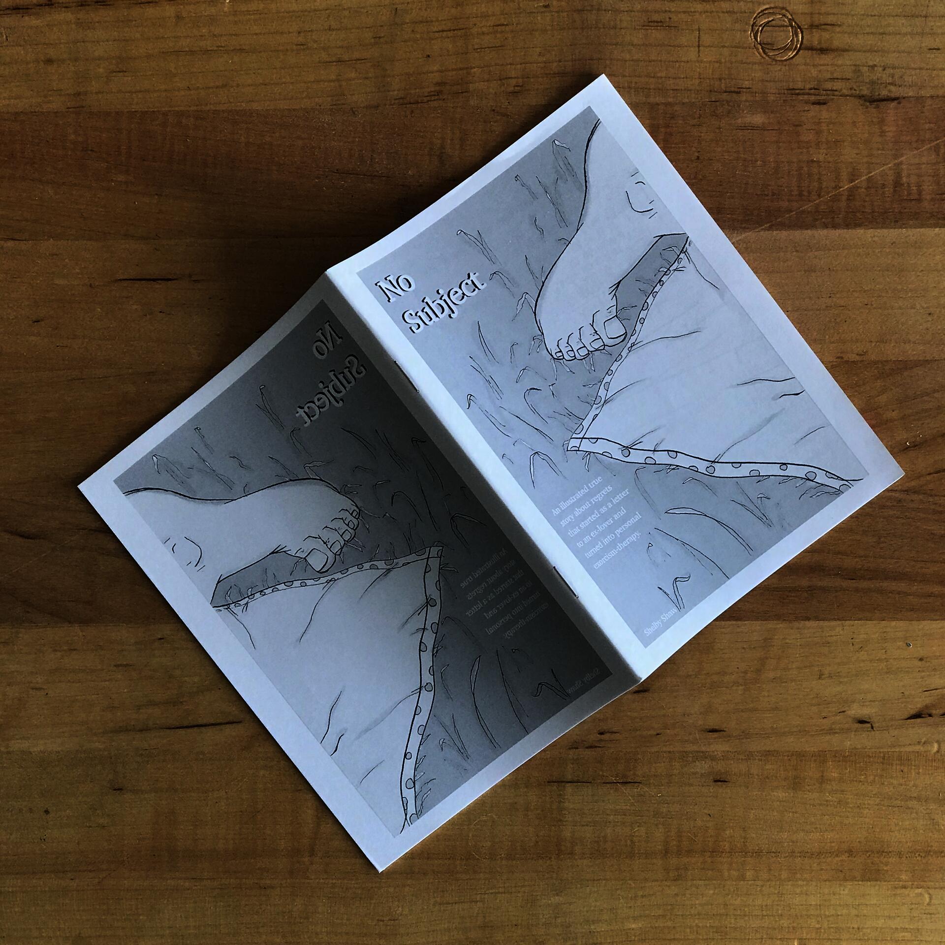 The book 'No Subject,' which has a black and white illustration of a foot in the grass on a blanket, is splayed open and lying pages-down on a wooden tabletop.