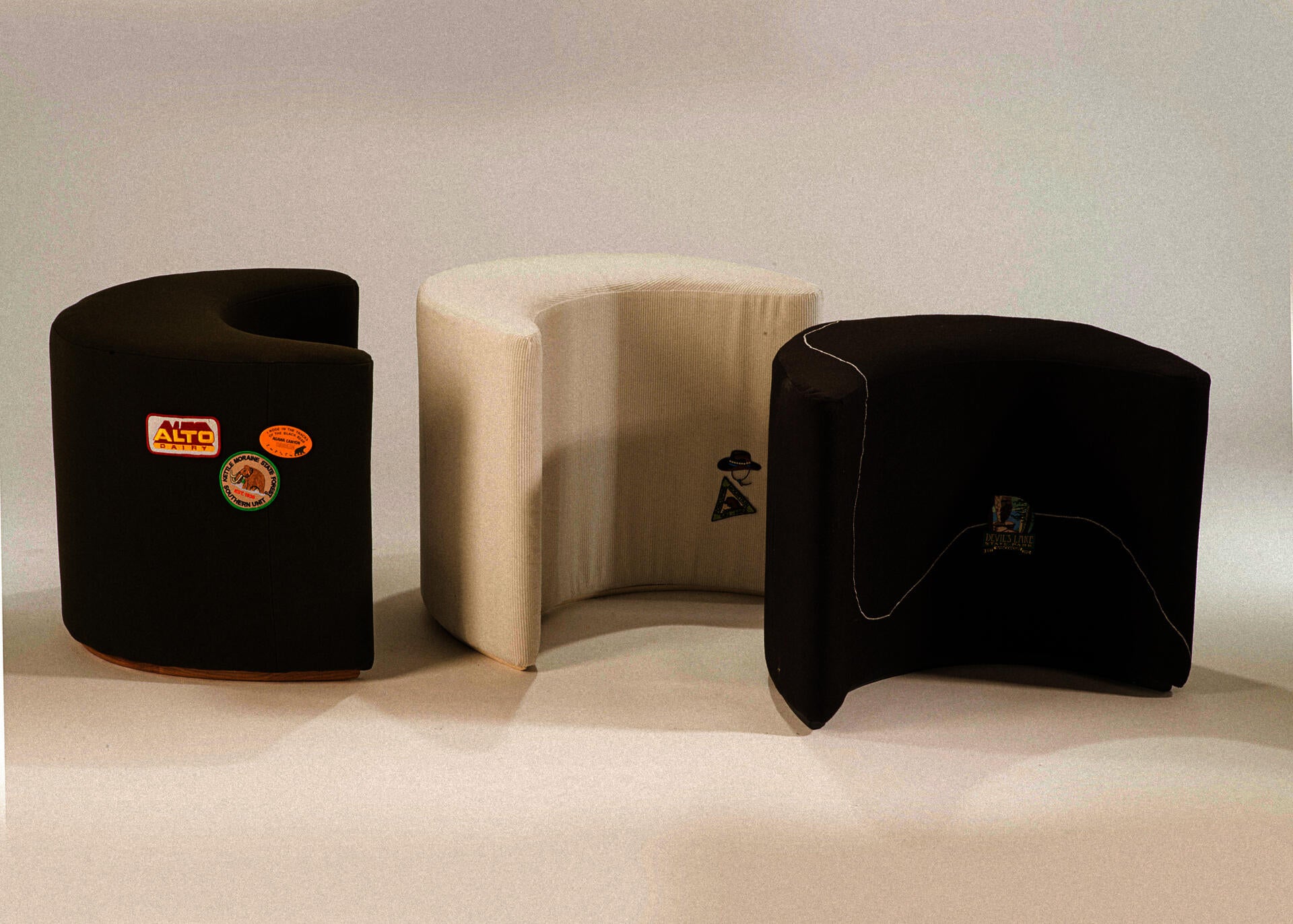 three crescent-moon shaped upholstered stools: one black, one deep green, and one white corduroy. embroidered patches are applied to the surface of all three.